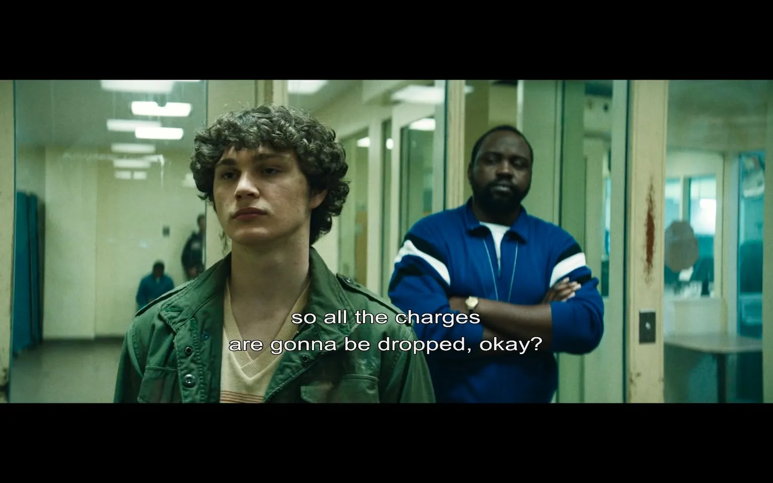 In this scene, two individuals are present in a stark, institutional setting that resembles a police station or detention center. The main subject is a young man with curly hair, appearing to be in his late teens or early twenties. He wears a green jacket over a light-colored sweater, and his expression is serious, suggesting concern or contemplation. He stands in front of a glass partition, facing another man who is slightly older and has a more robust build. This second individual is dressed in a blue tracksuit with white stripes and has his arms crossed, projecting a sense of authority or confidence.

The dialogue displayed on the screen reads, "so all the charges are gonna be dropped, okay?" indicating a discussion about legal matters, possibly a plea deal or negotiation. The background features muted colors and a few indistinct figures, hinting at a busy environment typical of a law enforcement facility. The overall atmosphere is tense, reflecting the gravity of the situation being discussed. There are no visible brands or logos, and the scene does not contain any elements of violence or sexual content.