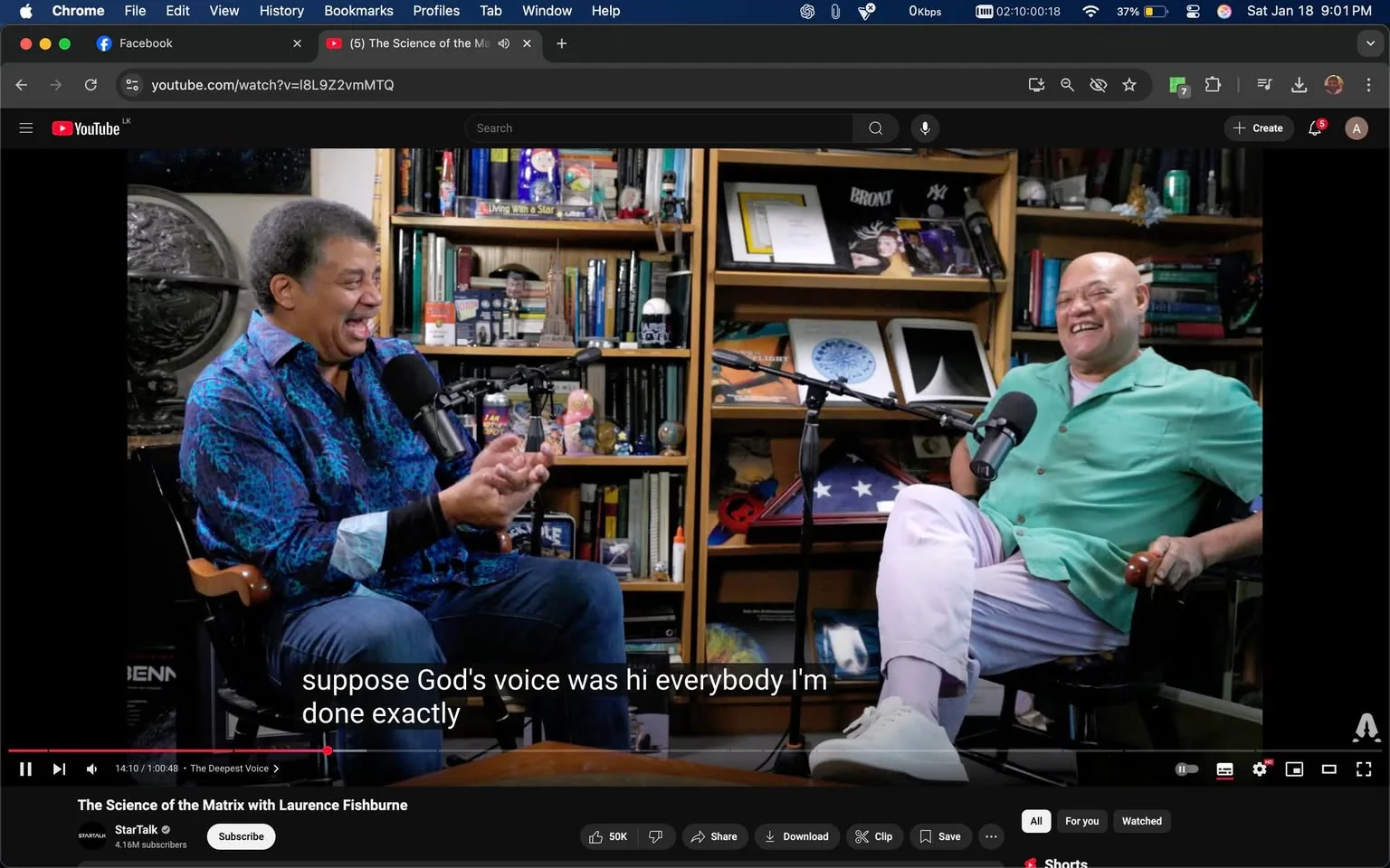 In the image, two unidentified adults are seated across from each other, engaged in a lively conversation. One individual, wearing a vibrant blue patterned shirt, is laughing and gesturing with his hands, suggesting a humorous or animated discussion. The other individual, dressed in a light green shirt and white pants, is also smiling, indicating enjoyment of the exchange. Both are positioned in front of microphones, suggesting a podcast or interview setting. The background features shelves filled with books, scientific models, and various decorative items, creating an intellectual atmosphere. The visible text at the bottom of the image appears to be a subtitle from the conversation, adding context to their discussion.