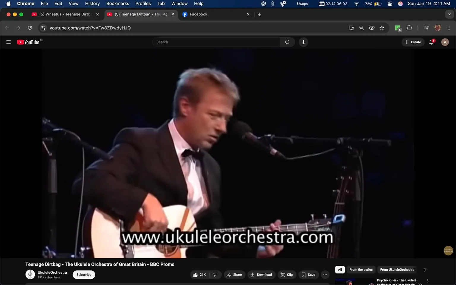 The image captures a live performance by a member of the Ukulele Orchestra of Great Britain, who is playing an acoustic guitar while seated. The musician is dressed in a formal black suit with a white shirt and a black bow tie, suggesting a polished and professional appearance suitable for a concert setting. He appears focused on his performance, with his head slightly bowed and his expression serious. 

In the background, there is a microphone positioned in front of him, indicating he is likely singing or speaking as he plays. The stage is dimly lit, creating an intimate atmosphere typical of live music events. The visible text on the screen includes the website URL "www.ukuleleorchestra.com," which is prominently displayed, suggesting this is a promotional video for the group. 

No signs of violence, sexual content, or any other sensitive material are present in the image. The overall setting appears to be a concert hall or theater, with a dark background that emphasizes the performer.