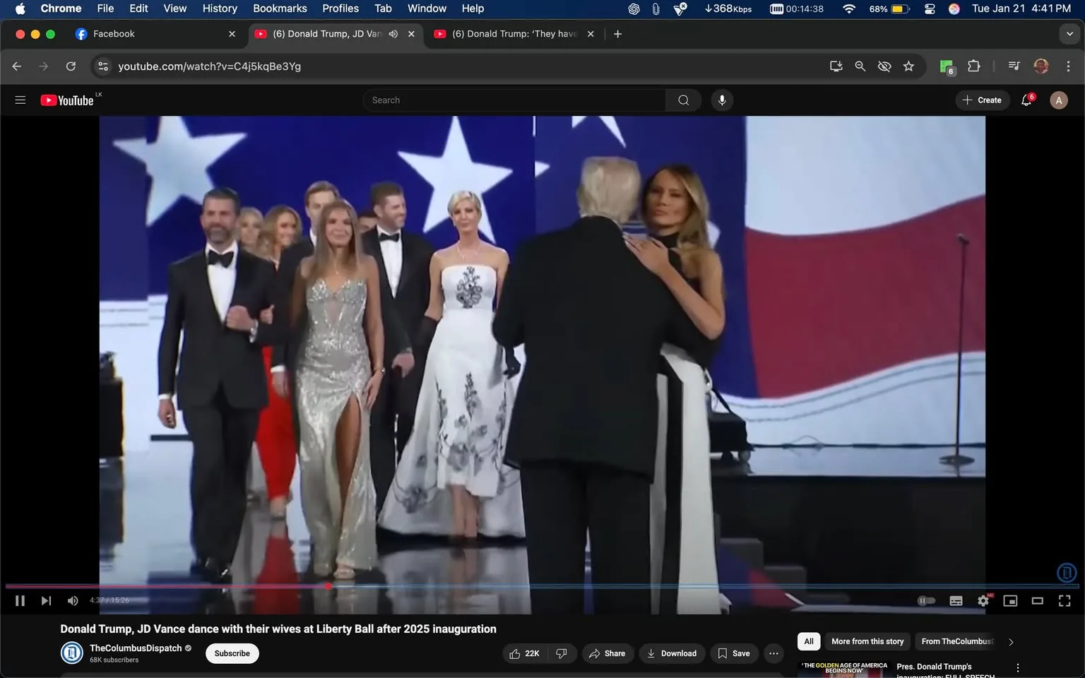 In the image, a group of individuals is gathered on a stage, likely at a formal event such as a ball or celebration. The main focus is on a man in a black suit, who appears to be engaging with a woman in a black and white dress, suggesting a moment of intimacy or celebration. The woman has long hair and is wearing a fitted dress that contrasts with the more traditional attire of the other attendees. 

To the left, a man in a tuxedo is walking with a woman in a sparkling silver gown that features a high slit, indicating a glamorous atmosphere. Other individuals in the background are dressed in formal wear, including tuxedos and elegant dresses, contributing to the upscale event vibe. The backdrop features large American flags and stars, reinforcing a patriotic theme. 

The setting appears to be indoors, with a well-lit stage and a polished floor, typical of formal gatherings. There are no visible logos or brands in the image, and the expressions of the individuals suggest a celebratory mood. The overall context hints at a political or celebratory event, possibly related to an inauguration or significant public gathering.