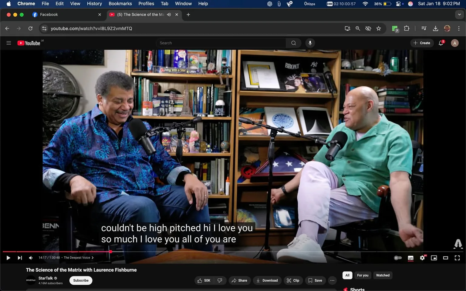 In the image, two men are seated across from each other, engaged in a lively conversation. One man, wearing a vibrant blue patterned shirt, appears to be laughing, showcasing a warm and friendly demeanor. He has short, curly hair and is positioned slightly to the left. The other man, dressed in a light green shirt and white pants, leans back in his chair, also smiling and seemingly enjoying the discussion. Both individuals are microphone-equipped, indicating they are likely recording a podcast or video. 

The setting is indoors, with a bookshelf filled with various books, objects, and decorations in the background, suggesting a casual yet intellectual atmosphere. Notable items on the shelves include what appears to be a globe and framed pictures, contributing to the scholarly vibe of the space. The lighting is bright, enhancing the cheerful mood of the interaction. There are no visible signs of violence, sexual content, or any brands in the image.