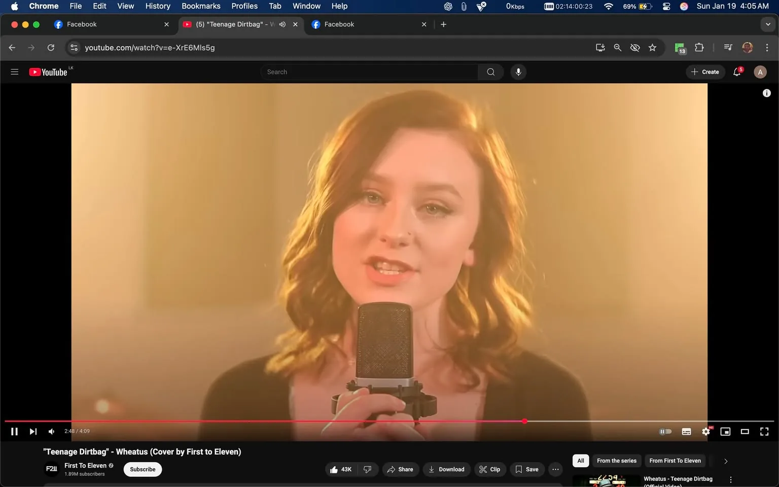 In the image, a young woman is singing into a microphone, appearing to be in a performance setting. She has medium-length, wavy brown hair and is wearing a black top. The lighting is warm and soft, creating a golden hue around her, which adds to the intimate atmosphere of the scene. The background is blurred, focusing attention on her expression and the microphone. She appears to be engaged and passionate about her performance, with a slight smile on her face. The video is titled "Teenage Dirtbag" - Wheatus (Cover by First to Eleven), indicating that this is a musical cover performance. The video interface shows that it is currently at 2:48 of a 4:09 duration, with options to like, share, and subscribe visible below. There are no visible brands or logos in the image, and the content does not contain any sexual or violent elements.