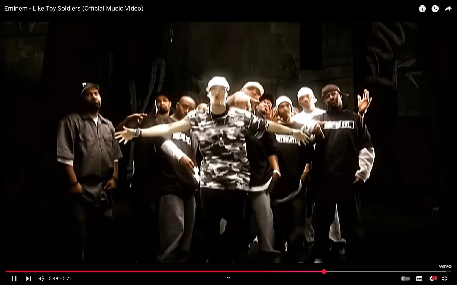 In this scene from the music video for "Like Toy Soldiers" by Eminem, the artist stands prominently in the center, arms outstretched, wearing a camouflage tank top and a white cap. He is surrounded by a group of men, all dressed in black shirts with white lettering, some wearing caps and bandanas. The expressions on their faces range from serious to engaged, creating a sense of camaraderie and intensity. The setting appears to be an indoor space with dark walls, possibly a warehouse or studio, illuminated by a spotlight that highlights the group. The background is minimal, focusing attention on the performers. The overall atmosphere conveys themes of unity and struggle, resonating with the song's lyrics about conflict and resilience. There are no visible brands or logos in the scene, and no elements of violence or sexual content are present.