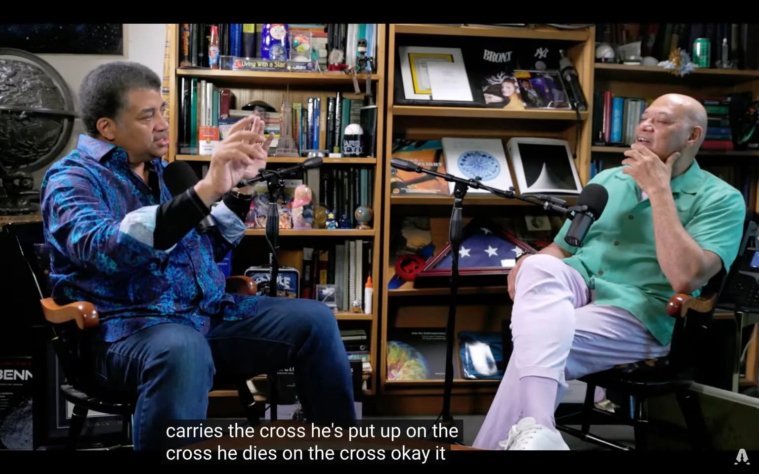 In the image, two individuals are seated in a cozy indoor setting, likely a studio or a personal office, surrounded by shelves filled with books, scientific models, and various decorative items. The person on the left, wearing a vibrant blue patterned shirt with a black undershirt, is animatedly gesturing with their hands while speaking. They appear to be engaged in a serious discussion, possibly about a philosophical or scientific topic. The individual on the right is dressed in a light green short-sleeved shirt and pale lavender pants, resting their chin on their hand, seemingly listening intently. 

Both individuals are positioned near microphones, indicating that this could be part of a podcast or interview setup. The background features a globe, books, and various knick-knacks, contributing to an intellectual atmosphere. There are no visible brands or logos in the image, and the overall tone is conversational and thoughtful. The lighting suggests it is an indoor setting, likely well-lit for video recording.