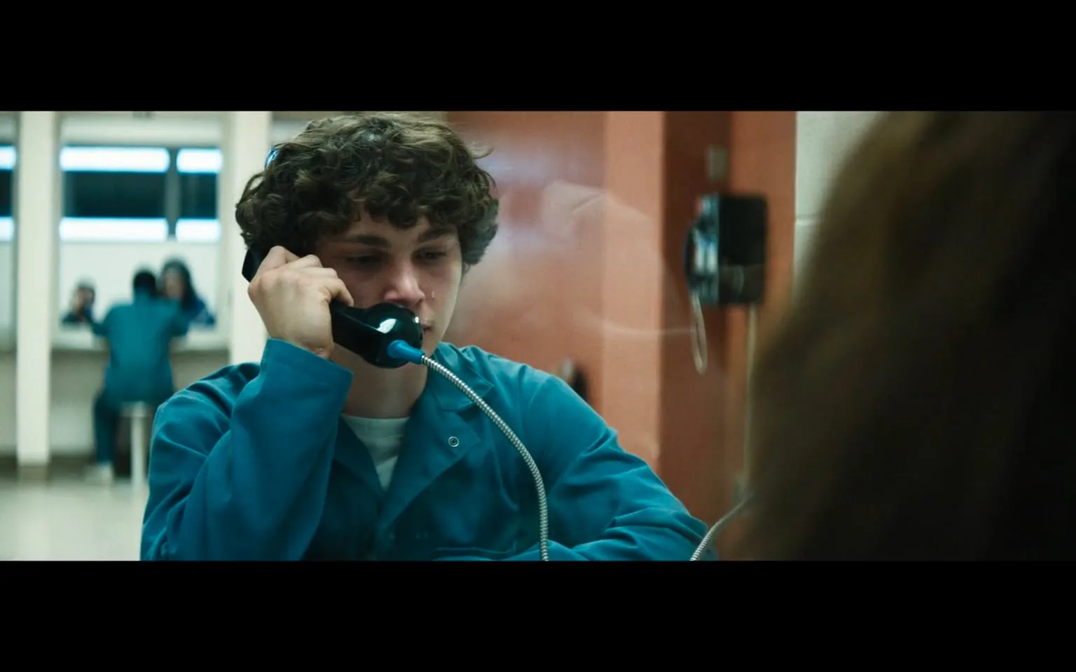 In the image, a young male, likely in his late teens, is seated at a phone booth, visibly distressed. He has curly hair and is wearing a blue, institutional-style outfit, which suggests he may be in a correctional or psychiatric facility. His expression is somber, with tears visible on his face, indicating he is experiencing a strong emotional moment. He holds a black telephone receiver to his ear, seemingly engaged in a serious conversation. 

In the background, two figures in similar blue outfits are seated at a table, their faces obscured, which adds to the somber atmosphere of the setting. The environment appears to be an indoor facility, with stark lighting and minimal decor, emphasizing the seriousness of the situation. The overall mood is tense and reflective, suggesting themes of isolation and emotional struggle.