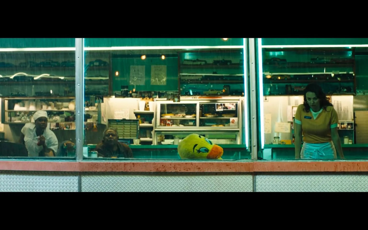 In the image, a diner scene is captured through a large window, showcasing a bustling yet intimate atmosphere. Two individuals are visible inside the diner: one is a man wearing a white chef's outfit, including a chef's hat, who appears to be engaged in conversation or observation, and the other is an older man seated nearby, looking out with a contemplative expression. Both seem to be in a relaxed setting, possibly during a quiet moment of the night.

In the foreground, a large, plush yellow duck head is resting on the counter, adding a whimsical touch to the scene. The woman in a yellow shirt and white apron stands nearby, leaning forward with her hands on the counter, suggesting she might be interacting with the customers or observing the situation. The diner is illuminated with soft, greenish lighting, creating a cozy ambiance, while the background reveals shelves filled with various items, including plates and small collectibles, hinting at a nostalgic diner theme.

The overall mood of the image is calm, with the rain visible on the window, suggesting it might be evening or night. There are no visible signs of violence, sexual content, or any explicit themes, making it a straightforward depiction of a diner setting.