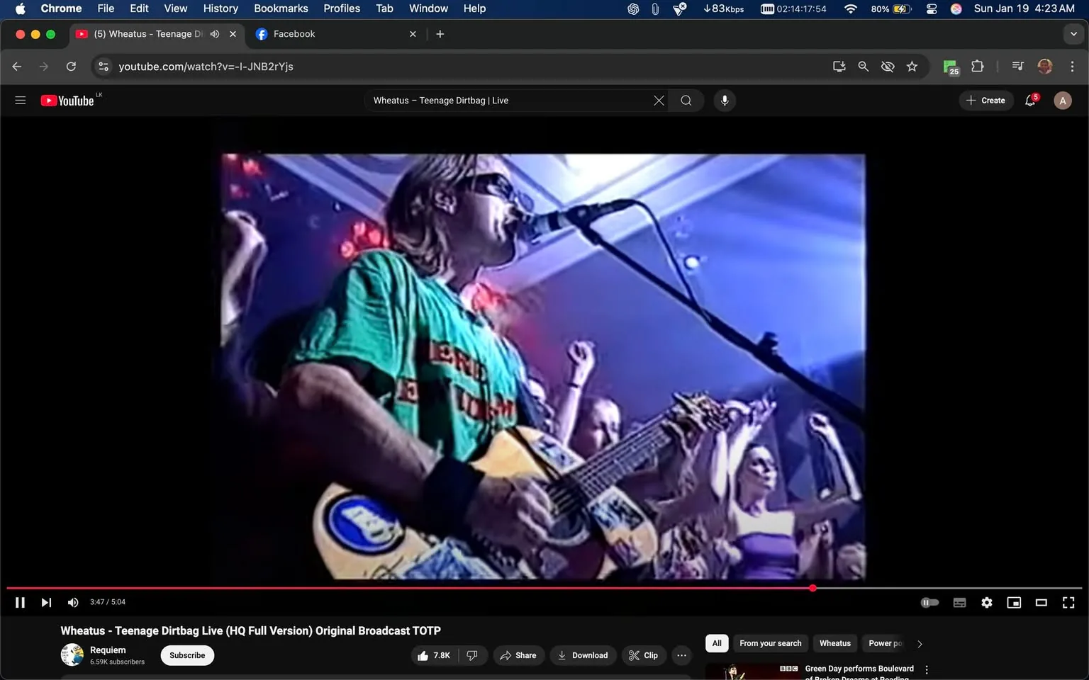 In the image, a musician is performing on stage, holding an acoustic guitar adorned with various stickers. He has long hair and is wearing a green t-shirt with red text, which appears to be a playful or humorous message. The setting is a lively indoor venue, likely during a concert, as indicated by the presence of bright stage lights and an enthusiastic audience. 

The audience members, visible in the background, are raising their arms, engaging with the performance. Some appear to be singing along, suggesting a high-energy atmosphere. The lighting is dynamic, with a mix of colors illuminating the scene, enhancing the excitement of the live event. The musician is focused on his performance, singing into a microphone, while the crowd's energy adds to the overall vibe of the moment. There are no visible signs of violence, sexual content, or any brands that can be identified in the image.