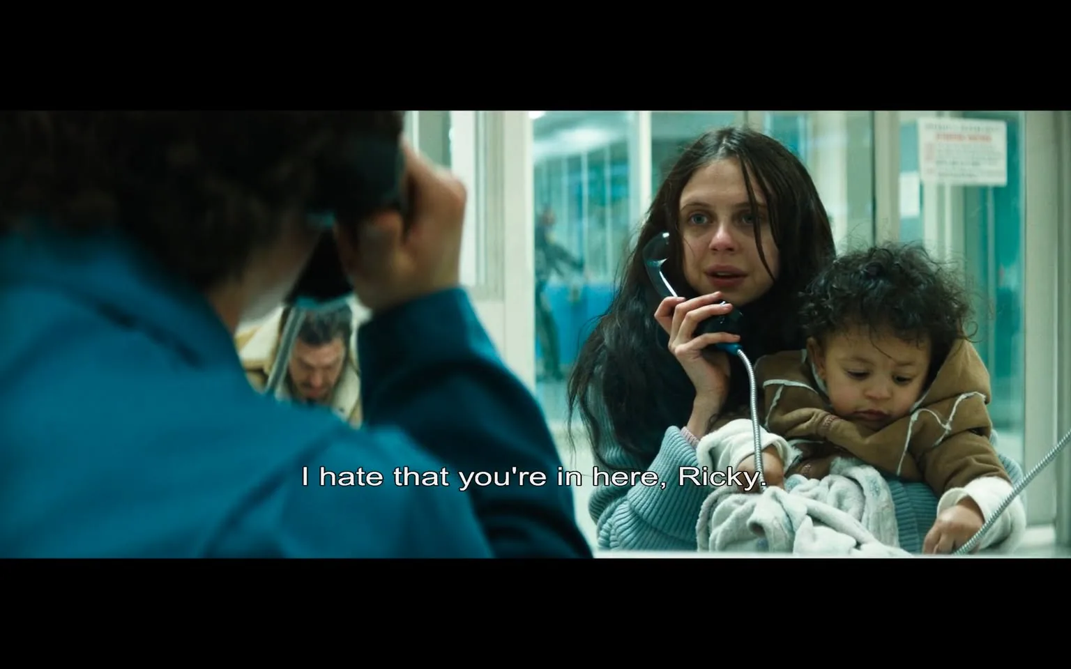 In the scene, a woman with long, dark hair appears distressed as she speaks on a telephone. She is holding a small child, who has curly hair and is dressed in a brown jacket. The woman is wearing a light blue sweater and has a pained expression, indicating emotional turmoil. The dialogue on the screen reads, "I hate that you're in here, Ricky," suggesting a conversation with someone incarcerated. 

In the background, there are blurred figures, including a man in a beige coat, which adds to the setting's somber atmosphere. The environment appears to be a visitation area, likely in a correctional facility, characterized by glass partitions and a clinical, institutional feel. The lighting is bright but harsh, contributing to the overall tension of the scene. The emotional weight of the moment is palpable, highlighting themes of separation and distress.