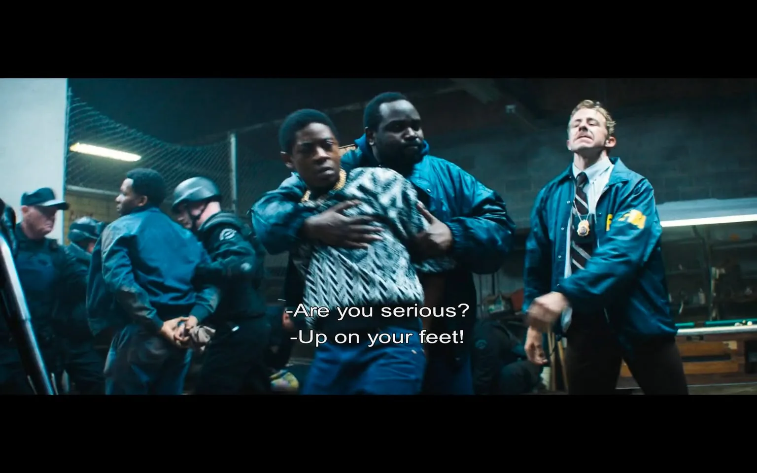 In a chaotic scene, a young man is being restrained by a larger individual, likely a law enforcement officer, who is holding him firmly. The young man appears distressed, wearing a patterned sweater and a chain around his neck, while the officer is dressed in a blue jacket. The background reveals several other officers in tactical gear, suggesting a high-stress situation, possibly an arrest or intervention. The setting is indoors, with a concrete wall and a chain-link fence visible, indicating a police or holding area. 

The dialogue suggests urgency and frustration, with one officer asking, "Are you serious?" and another commanding, "Up on your feet!" This indicates a confrontational atmosphere. The expressions of the individuals involved convey tension, with the young man looking apprehensive and the officers appearing assertive. The overall mood is intense, reflecting a moment of conflict or authority being exerted. There are no visible brands or logos in the scene, and the focus remains on the interaction between the officers and the detained individual.