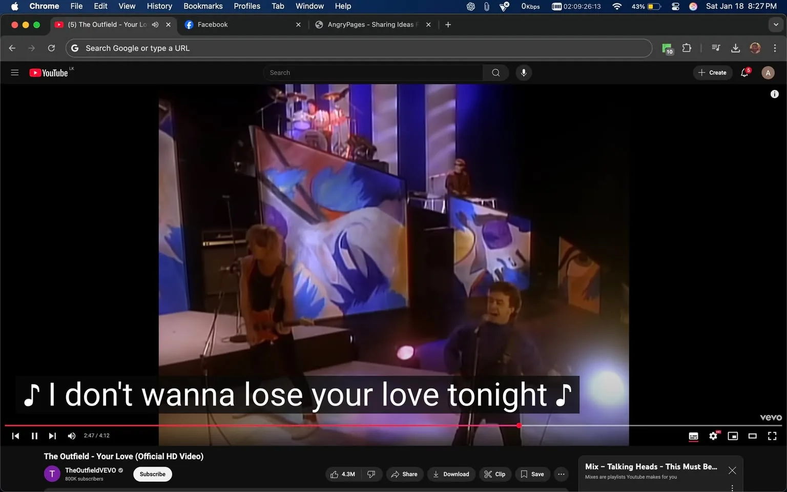 The scene captures a performance from a music video featuring a band. In the foreground, an unidentified adult is playing an electric guitar while wearing a black tank top and white sneakers. Another unidentified adult is singing into a microphone, dressed in a blue outfit. The background displays large, colorful abstract panels with blue and purple designs, creating a vibrant stage setting. The lighting is dynamic, with soft pink and blue hues illuminating the performers. Lyrics are visible on the screen, stating, "I don't wanna lose your love tonight," indicating the song being performed. The overall atmosphere suggests a lively musical event.