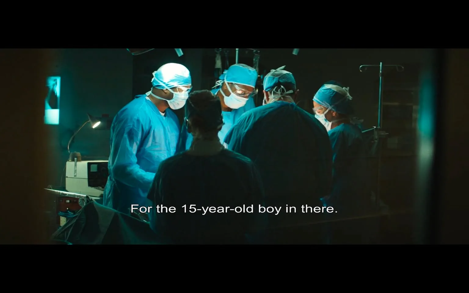 In the image, a group of five medical professionals is gathered around a surgical table, focused on a procedure. They are dressed in blue surgical scrubs and masks, indicating a sterile environment typical of an operating room. The lighting is dim, with a bright light illuminating the area where they are working. The professionals appear to be engaged in a serious discussion, with one of them speaking the line, "For the 15-year-old boy in there," suggesting the urgency and importance of the situation. 

The background features medical equipment, including an IV stand and monitors, which are common in surgical settings. There are no visible logos or brands in the image. The overall atmosphere conveys a sense of tension and professionalism, characteristic of a critical medical operation. The focus is clearly on the teamwork and dedication of the medical staff as they work to save a young patient.
