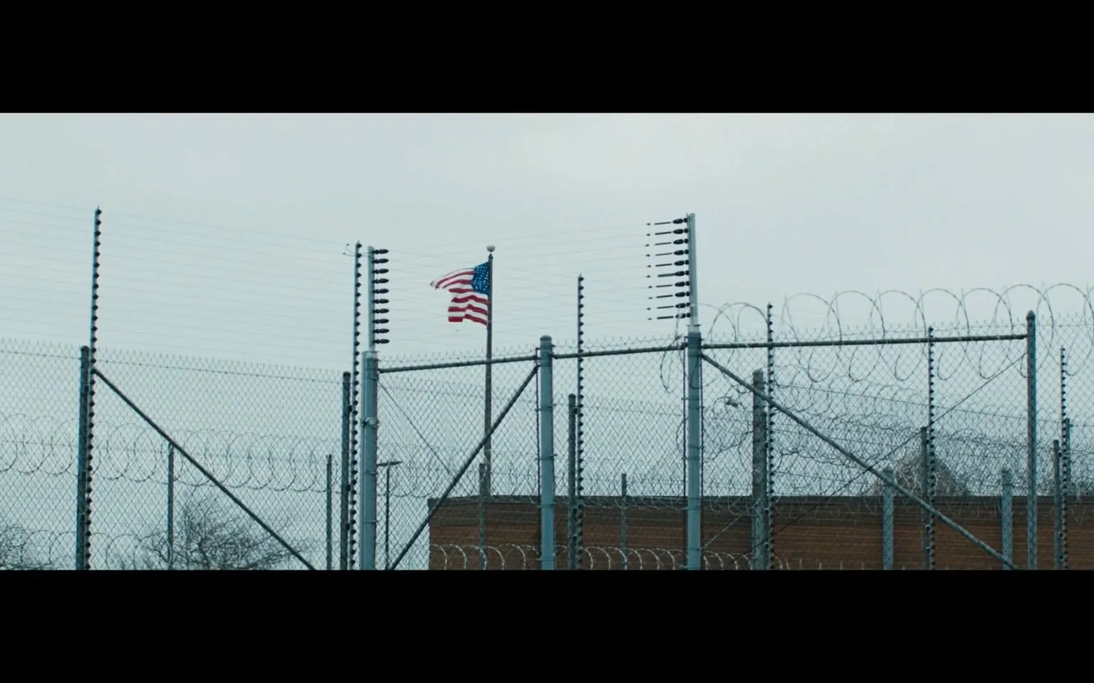 The scene captures a stark prison environment, characterized by high chain-link fences topped with barbed wire. In the background, an American flag flutters gently, indicating a somber atmosphere. The sky is overcast, contributing to the bleakness of the setting. The fences are equipped with security features, suggesting a high level of surveillance and control. The overall impression is one of confinement and restriction, typical of correctional facilities. No people or additional objects are visible in the frame, emphasizing the isolation of the location.