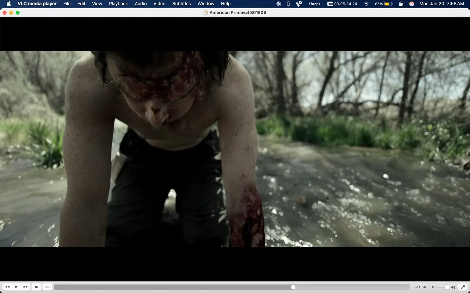 In the image, a male figure is seen crawling on the ground, appearing to be in distress. He is shirtless, revealing a torso that is smeared with blood, particularly noticeable on his arms and forehead. The blood suggests he has sustained injuries, contributing to a sense of violence in the scene. His expression is not fully visible, but the posture indicates a struggle or desperation. The setting appears to be outdoors, near a shallow stream or body of water, with blurred greenery in the background, hinting at a natural environment. The lighting suggests it is daytime, possibly in a rural or wilderness area. The overall atmosphere is tense and unsettling, emphasizing the character's plight.