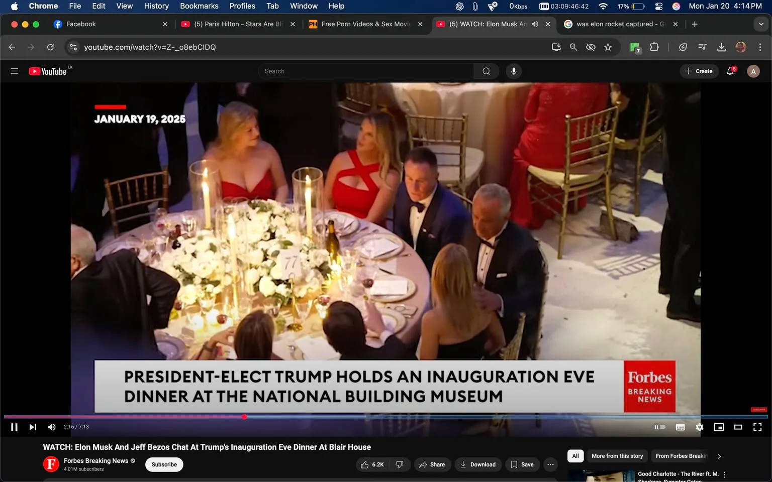 The scene captures a formal dinner event, likely an inauguration eve gathering, set at the National Building Museum. The table is elegantly decorated with a floral centerpiece featuring white roses and lit candles, creating a warm ambiance. Attendees are dressed in formal attire; several women are wearing striking red dresses, while men are in tuxedos. Notably, one woman in a red dress has a distinctive cut-out design, and another is seated nearby in a more traditional gown. 

The atmosphere appears lively, with guests engaged in conversation. The table setting includes fine china, glassware, and bottles of wine, suggesting a high-end dining experience. A placard with the number "77" is visible on the table, possibly indicating the table number for the event. 

In the background, other guests can be seen, and the overall setting is indoors, illuminated by soft lighting, indicative of an evening event. The visible text at the bottom of the image references the event as a dinner held by the president-elect, adding a political context to the gathering.
