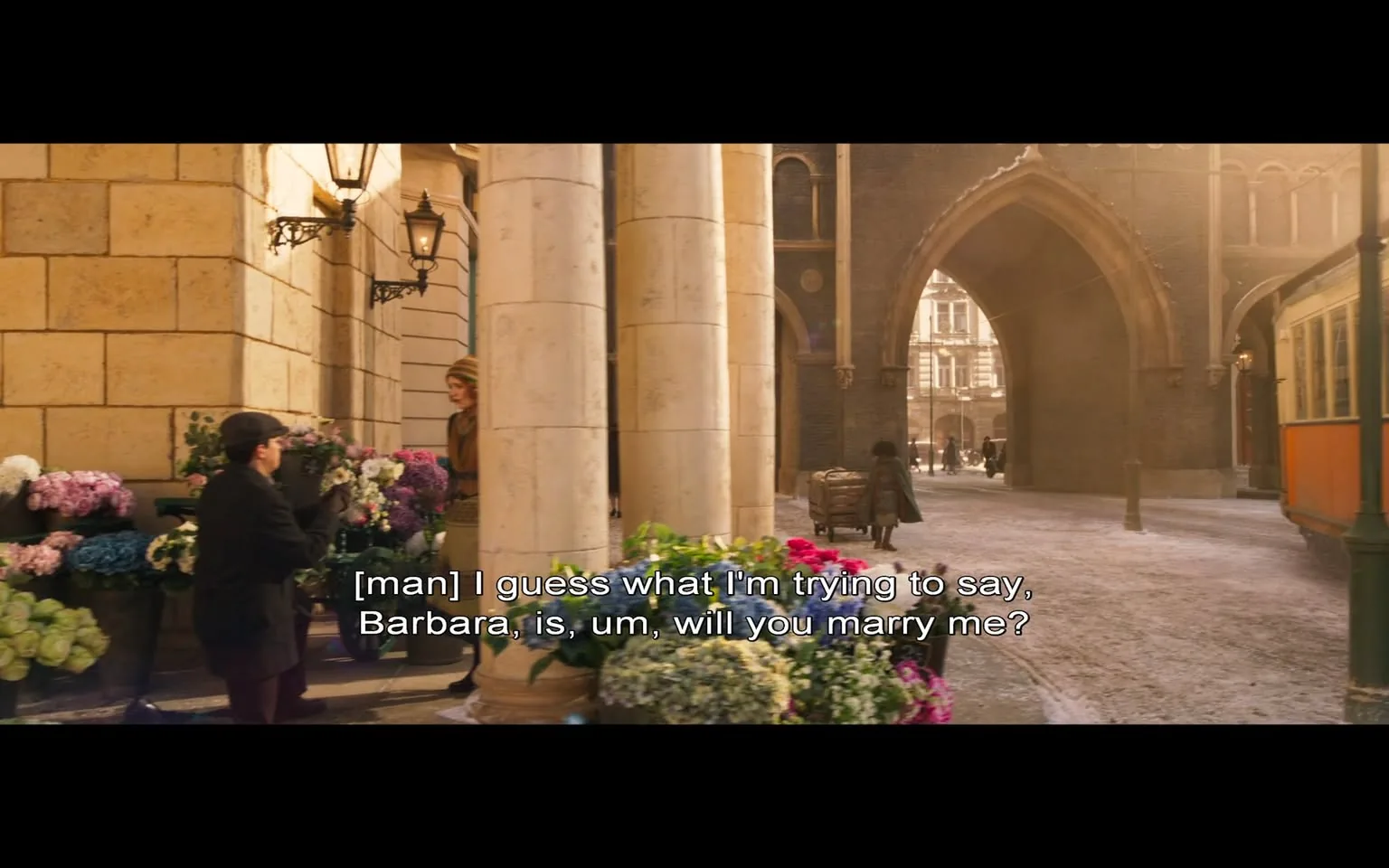 In this scene, a man is kneeling in front of a woman, seemingly in a moment of emotional significance, as he proposes marriage. The man is dressed in a dark coat and a cap, while the woman stands nearby, wearing a stylish outfit with a hat. The setting appears to be an outdoor market or street, characterized by stone architecture and decorative lampposts. Flower stalls filled with vibrant blooms, including pink and blue flowers, line the area, adding color to the scene. 

The background features an archway leading to another street, where a few pedestrians can be seen walking. The lighting suggests it is either early morning or late afternoon, creating a warm, inviting atmosphere. The text overlay indicates the man is expressing his feelings, stating, "I guess what I'm trying to say, Barbara, is, um, will you marry me?" This adds a layer of intimacy and significance to the moment, highlighting the romantic theme of the scene. There are no visible brands or logos, and the overall mood is one of hope and anticipation.