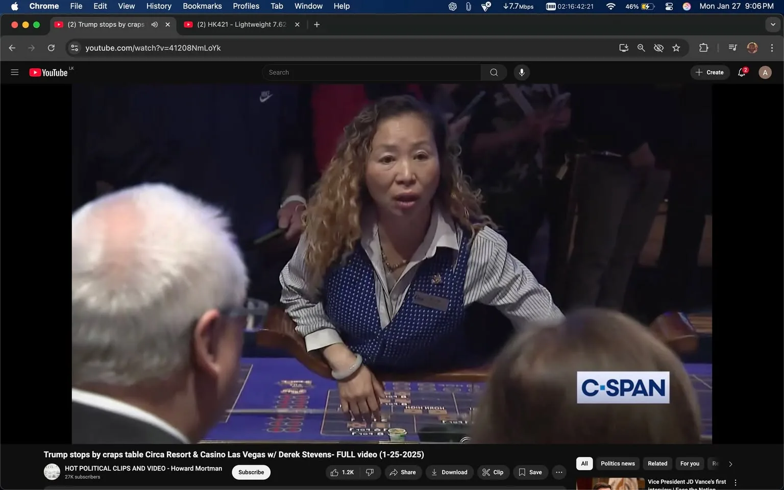 In a lively casino setting, a female dealer with curly hair and a friendly expression is engaged with players at a craps table. She wears a blue vest over a white shirt, which is typical attire for casino staff. The table is adorned with a blue felt surface featuring various betting areas and markings associated with the game of craps. The dealer appears to be explaining something or responding to a question, leaning slightly forward with her hands on the table. 

In the background, several people are gathered around the table, some focused on the game while others seem to be using their phones. The atmosphere is bustling, indicative of a busy casino environment, likely during the evening. The lighting is bright, typical of indoor casino settings, enhancing the vibrant colors of the table and the players' attire. No visible brands or logos are present in the image. The scene captures the excitement and social interaction characteristic of a casino gaming experience.