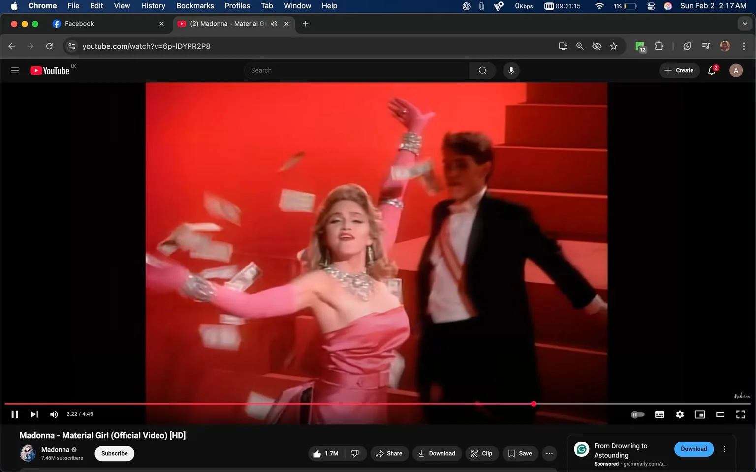 In this vibrant scene from a music video, a woman dressed in a glamorous pink gown is the focal point. She has long, wavy blonde hair and is adorned with sparkling jewelry, including a necklace and bracelets. Her left arm is raised dramatically, and she appears to be joyfully tossing what looks like dollar bills into the air, creating a sense of celebration and abundance. The background is a bold red, which enhances the lively atmosphere of the scene. 

Beside her, a man in a black tuxedo with a white shirt and a red sash is present, moving slightly behind her. His expression is less prominent, but he seems to be engaged in the performance. The setting appears to be a stage, indicated by the steps leading up in the background. The overall mood is energetic and theatrical, typical of a pop music performance. There are no visible logos or brands in the image, and the content does not contain any sexual or violent elements.