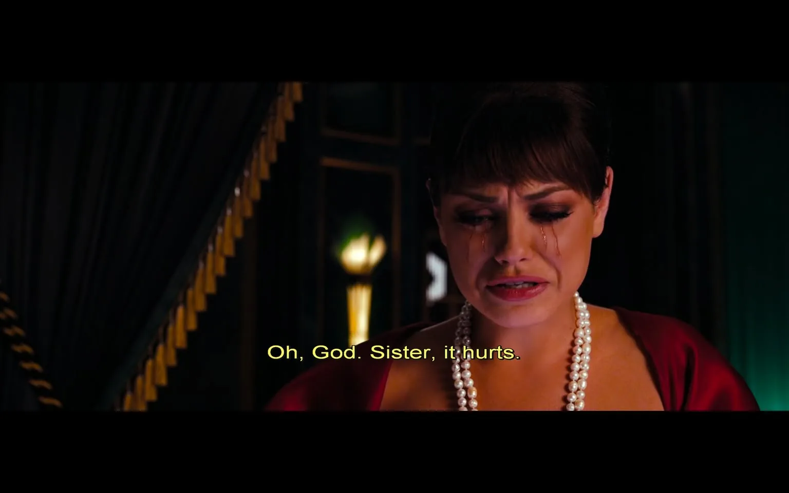 In the scene, a woman is visibly distressed, with tears streaming down her face, indicating a moment of emotional turmoil. She is wearing a deep red dress that contrasts with her fair skin, and she has a string of pearls around her neck, adding a touch of elegance to her appearance. Her hair is styled in a neat updo, and her makeup is dramatic, with dark eyeliner accentuating her eyes. The setting appears to be indoors, with dark curtains in the background and soft lighting that creates a somber atmosphere. The text overlay reads, "Oh, God. Sister, it hurts," suggesting a conversation with someone off-screen, possibly a sibling, indicating a personal and emotional moment. The overall mood of the image is one of sadness and vulnerability, reflecting a significant emotional struggle.