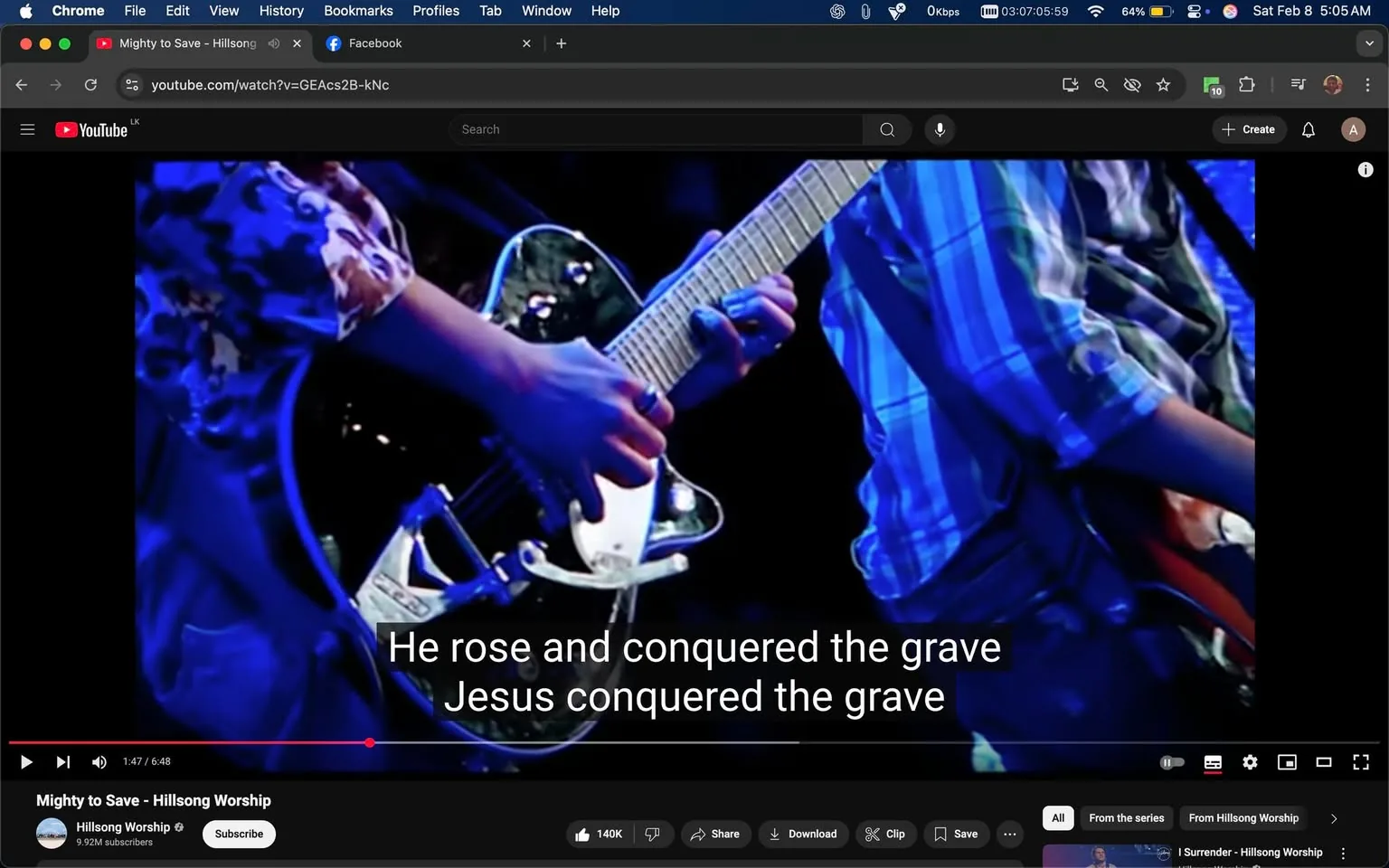 The image shows a close-up of two musicians performing on stage, likely during a worship service. One musician is playing an electric guitar, which has a shiny black body and a white pickguard. The hand on the guitar is adorned with a ring, and the musician is wearing a patterned shirt with floral designs. The other musician, positioned slightly behind, is playing a guitar with a lighter color and is wearing a blue and white plaid shirt. 

The background is illuminated with blue lighting, creating a vibrant atmosphere typical of live performances. The lyrics "He rose and conquered the grave Jesus conquered the grave" are displayed prominently on the screen, indicating that this is a worship song, possibly from Hillsong Worship, as suggested by the branding visible in the video player interface. The setting appears to be indoors, likely in a church or concert venue, and the time of day is not explicitly indicated. There are no visible signs of violence, sexual content, or any brands other than Hillsong.