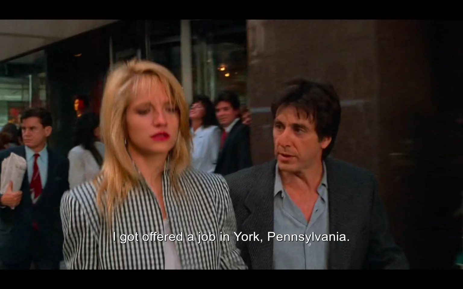 In this scene, two characters are walking through a bustling city street, likely in New York, given the urban setting. The woman, with long blonde hair, is wearing a black and white striped blazer, looking somewhat distressed or preoccupied. Her makeup includes bold red lipstick, and she appears to be in her late twenties or early thirties. The man beside her has dark hair and is dressed in a gray blazer over a light blue shirt, looking intently at her as they converse. 

The background is filled with pedestrians in business attire, suggesting a busy workday atmosphere. Some individuals are carrying newspapers, and there are blurred figures indicating movement, which adds to the sense of a lively urban environment. The dialogue visible on the screen indicates that the woman has received a job offer in York, Pennsylvania, hinting at a significant life decision. The overall mood of the scene seems to convey tension or concern regarding their conversation.