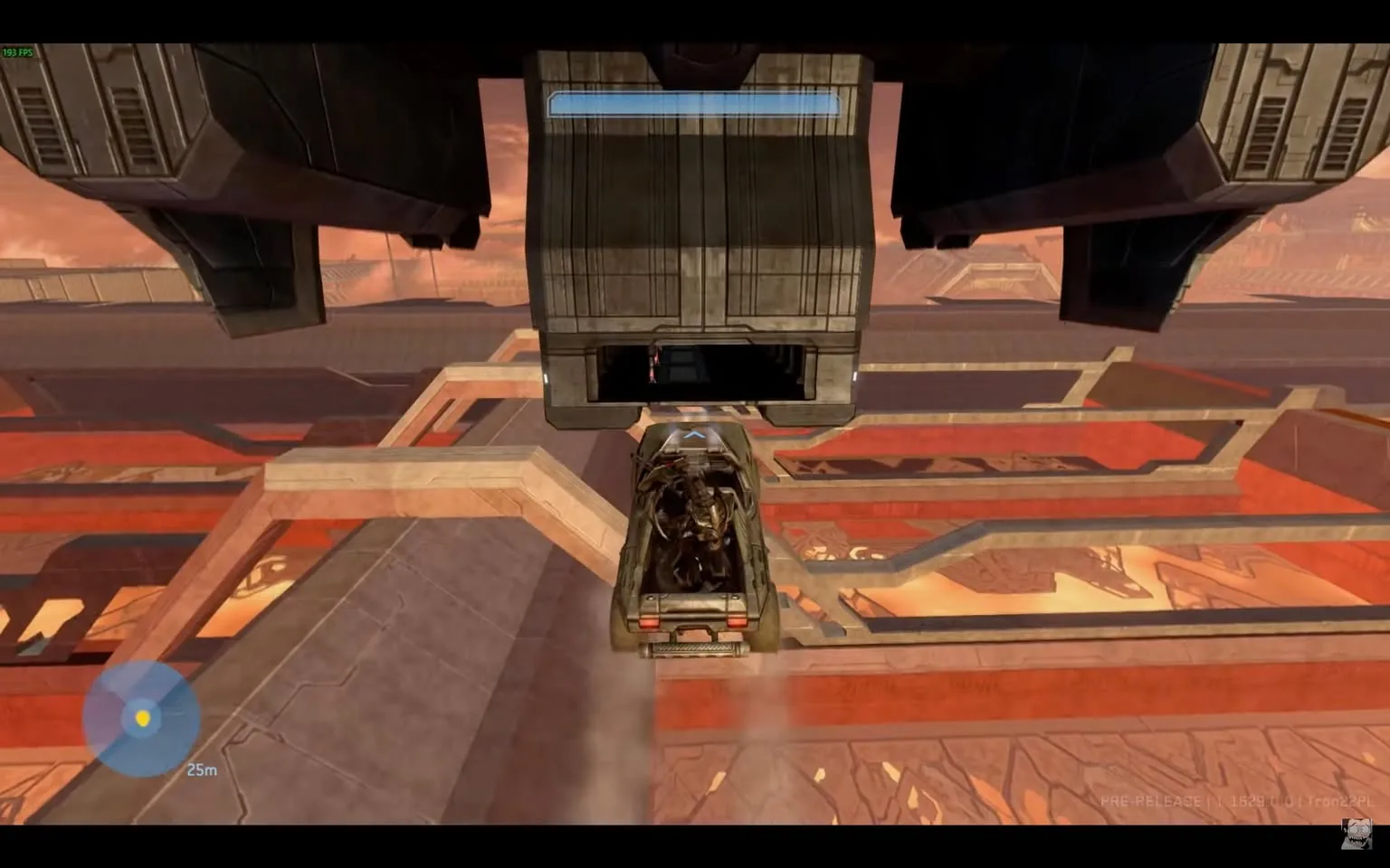 In the image, a vehicle is positioned directly beneath a large, hovering structure, likely part of a futuristic or sci-fi setting. The vehicle appears to be a military-style transport, possibly resembling those found in video games. It is being lifted by the structure above, which has a metallic, industrial design. The background features a complex landscape with geometric patterns and a mix of colors, predominantly shades of red and gray, suggesting a technologically advanced environment. The scene is illuminated by a soft light, hinting at either dawn or dusk. There are no visible characters or people in the image, focusing solely on the vehicle and the structure. The overall atmosphere conveys a sense of action or movement, typical of a gaming scenario.