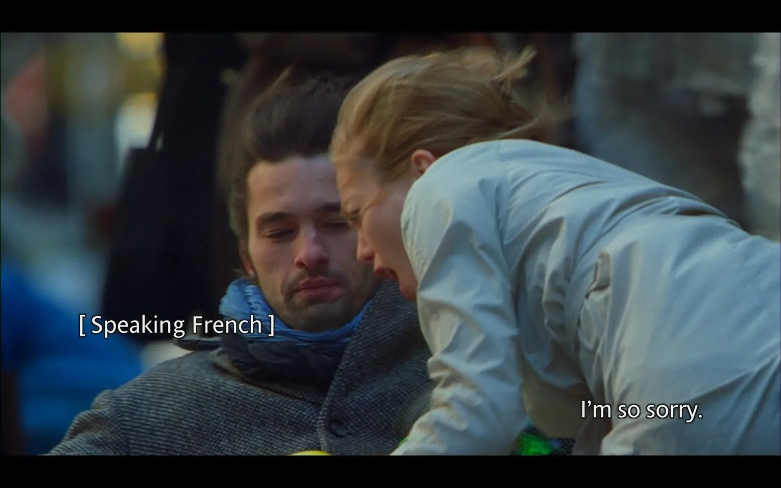 In the image, two individuals are present in a moment of emotional interaction. One person, a man, has a somber expression and appears to be listening intently, while the other, a woman, leans towards him, visibly distressed. She has her hair down and is wearing a light-colored jacket. The man is dressed in a dark coat with a blue scarf wrapped around his neck. The background suggests an outdoor setting, possibly in a busy urban area, with blurred figures and colors indicating movement and activity. 

Text overlays indicate that the man is speaking in French, while the woman expresses an apology, saying, "I'm so sorry." The scene conveys a sense of urgency and emotional weight, as the woman appears to be trying to convey something important to the man. There are no visible brands, logos, or indications of violence or sexual content in the image. The focus remains on the interpersonal dynamics between the two characters.