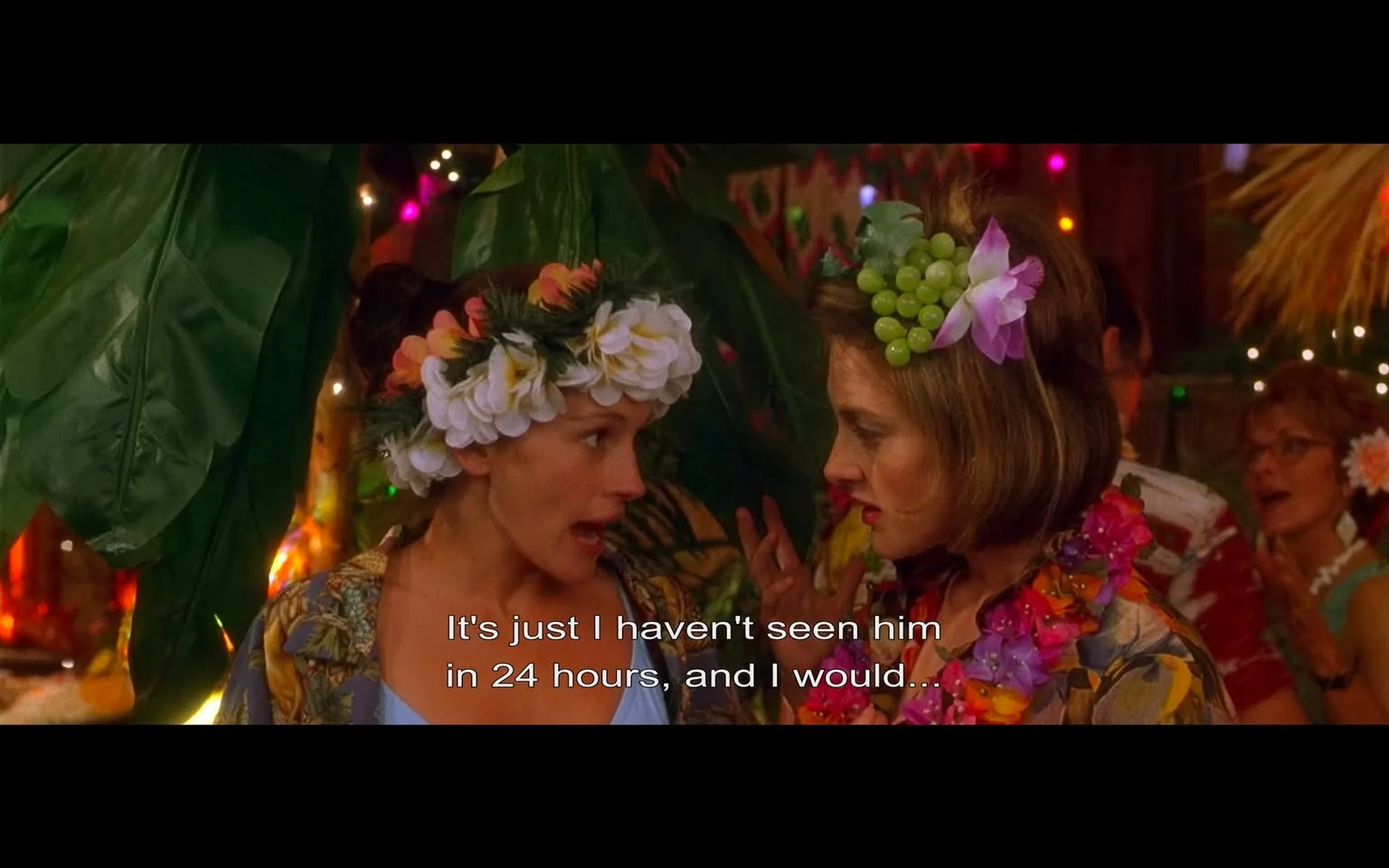 The image depicts a scene from a festive gathering, likely a tropical-themed party. 

**Readable Text:**
- The visible text at the bottom of the image reads: "It's just I haven't seen him in 24 hours, and I would..."

**People:**
- There are two main individuals in the foreground:
  1. The woman on the left has long, dark hair and is wearing a floral-patterned shirt. She has a floral crown made of white and pink flowers adorning her head. Her expression appears animated, suggesting she is engaged in a conversation.
  2. The woman on the right has shoulder-length brown hair and is wearing a similar floral-patterned shirt. She has a headpiece that includes green grapes and a purple flower. Her expression is serious as she listens to the woman on the left.

- In the background, there are additional party attendees, some of whom are partially visible. They are dressed in colorful, tropical attire, and some are wearing leis, contributing to the festive atmosphere.

**Clothing:**
- Both women are dressed in casual, tropical-themed clothing, featuring vibrant colors and floral patterns. The woman on the left has a shirt that is predominantly dark with colorful flowers, while the woman on the right's shirt has a lighter base with floral designs.

**Setting:**
- The background is decorated with large green leaves and colorful lights, creating a lively and festive ambiance. There are hints of a tropical or beach-themed decor, with elements like tiki torches or bamboo structures visible. The lighting is warm and inviting, suggesting an evening event.

- The overall environment appears to be a party setting, likely indoors, given the decorations and the presence of multiple people.

**Objects:**
- The image does not prominently feature any specific brands or products. The focus is primarily on the individuals and the festive decorations.

In summary, the image captures a vibrant and lively moment at a tropical-themed gathering, with two women engaged in conversation amidst a colorful and festive backdrop.