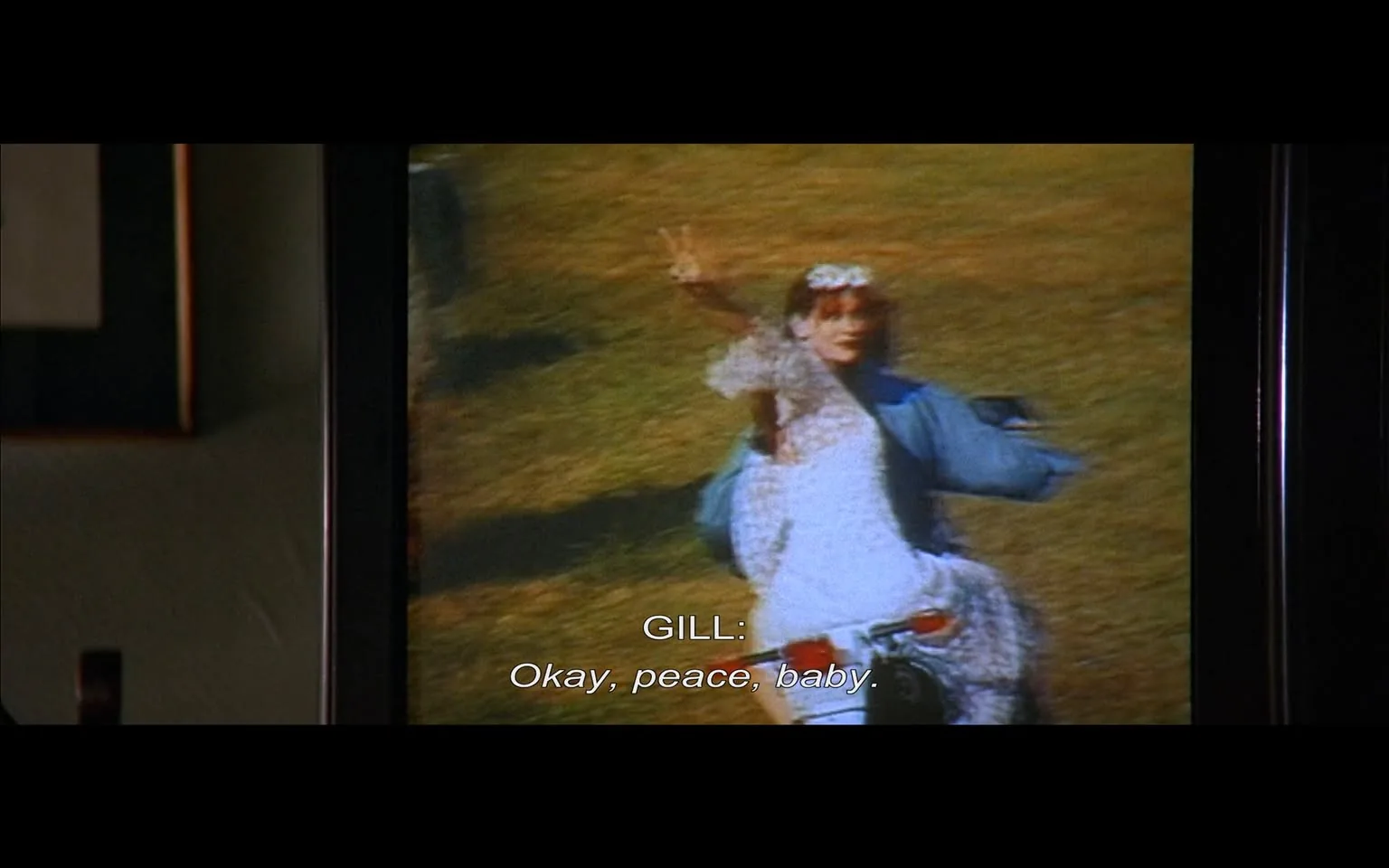 The image displays a scene from a film, captured on a television screen. The screen shows a slightly blurred and grainy image of a person, possibly a woman, who appears to be outdoors in a grassy area. She is wearing a white dress, which has a flowing design, and a flower crown on her head. The dress is adorned with lace or similar decorative elements. The individual is making a peace sign with her right hand, suggesting a carefree or celebratory mood.

The person is also wearing a blue jacket over the dress, which contrasts with the white fabric. The background features a green field, indicating an outdoor setting, possibly a festival or gathering. The colors in the image are somewhat muted, likely due to the quality of the television display.

Beneath the image, there is readable text that indicates a character named "GILL" is speaking. The dialogue reads: "Okay, peace, baby." This suggests a casual or informal tone in the conversation.

To the left of the television screen, there is a dark frame, possibly part of the television stand or wall decor, which includes a picture frame. The frame is rectangular and has a thin border, but the contents of the frame are not visible in this image.

Overall, the scene captures a moment of lightheartedness, with the focus on the individual in the dress and the dialogue suggesting a relaxed atmosphere. The image is framed within a larger context of a room, indicated by the presence of the wall and the picture frame.