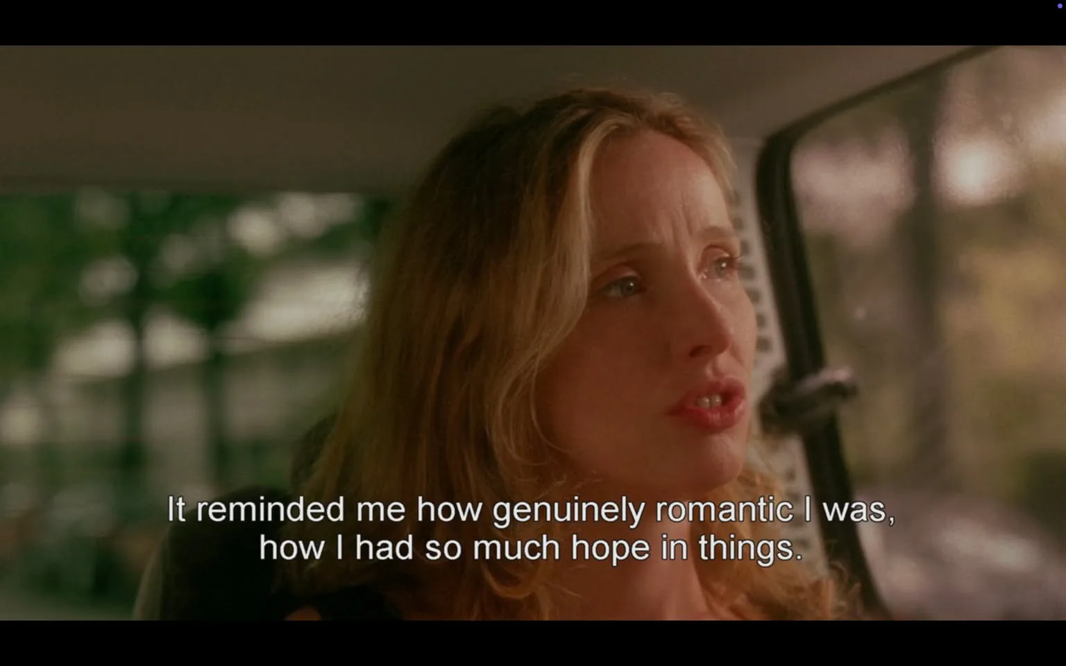 The image depicts a woman with long, wavy hair, seated in a vehicle. She appears to be in a moment of emotional expression, as indicated by her slightly parted lips and intense gaze. The background shows a blurred view of greenery, suggesting that the vehicle is in motion or parked in a natural setting. The lighting in the image has a warm, soft quality, contributing to the overall mood.

The woman is wearing a black top, which contrasts with her light-colored hair. Her facial expression conveys a sense of contemplation or nostalgia, as she seems to be reflecting on a personal experience. The text overlay at the bottom of the image reads: "It reminded me how genuinely romantic I was, how I had so much hope in things." This text is presented in a white font, making it easily readable against the background.

The interior of the vehicle is partially visible, with a hint of the dashboard and a side window. The window shows a blurred view of the outside, reinforcing the idea of movement. The overall composition of the image focuses on the woman's face and expression, with the text providing context to her thoughts.

No logos, brands, or identifiable public figures are present in the image. The setting appears to be casual and intimate, likely capturing a moment of personal reflection.