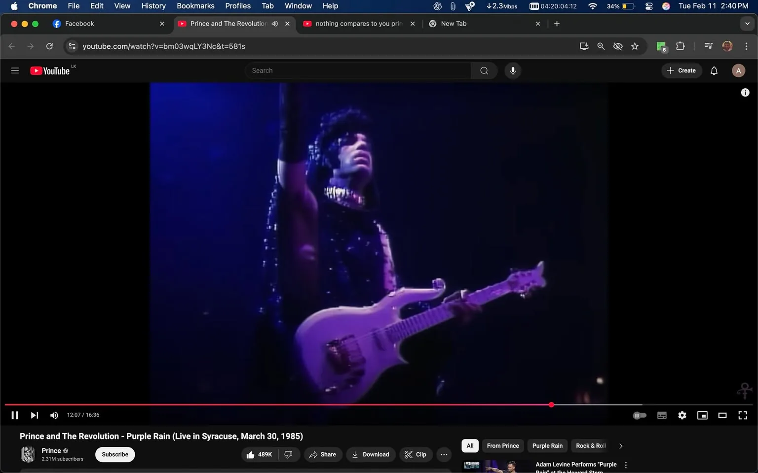 The image captures a scene from a live performance, specifically from a video titled "Prince and The Revolution - Purple Rain (Live in Syracuse, March 30, 1985)." 

In the foreground, a male figure is prominently featured, holding a white electric guitar. He is dressed in a black outfit adorned with sequins or sparkles, which reflects the stage lights, creating a shimmering effect. The outfit appears to be sleeveless, showcasing his arms. His hair is styled in a voluminous, curly manner, and he has a serious expression on his face, suggesting he is deeply engaged in the performance. 

The background is illuminated with blue and purple stage lights, contributing to a dramatic atmosphere. The lighting creates a contrast between the performer and the backdrop, enhancing the visual impact of the scene. 

The video player interface is visible at the bottom of the image, displaying the title of the performance, the duration (12:07 out of 16:36), and the number of subscribers to the channel (2.31M). There are also options for sharing, downloading, and other interactive features typical of a YouTube video player.

No additional text, logos, or trademarks are visible in the image aside from the YouTube interface elements. The overall setting indicates a concert or live music event, characterized by the performance of a well-known artist.