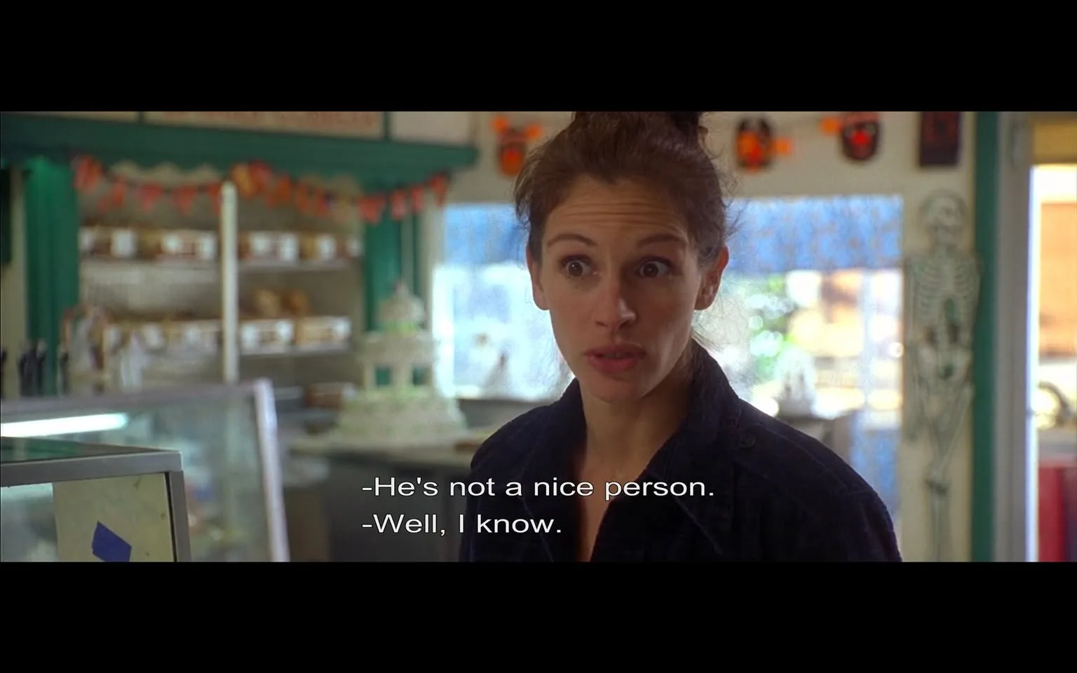 The image depicts a scene from a film set in a bakery or café. The interior features a colorful and inviting atmosphere, with green walls and decorations that include a garland of orange and black items, likely related to a holiday theme. In the background, there are shelves displaying various baked goods, including what appears to be bread and pastries. A white cake with decorative elements is prominently placed on a counter.

In the foreground, a woman is engaged in conversation. She has long, brown hair pulled back into a loose bun and is wearing a dark, button-up shirt. Her expression is one of concern or intensity, with wide eyes and slightly furrowed brows, suggesting she is involved in a serious discussion. The dialogue displayed on the screen indicates a conversation about someone's character, with one person stating, "He's not a nice person," to which the woman responds, "Well, I know."

The setting is well-lit, with natural light coming from a window in the background, which adds to the warm ambiance of the space. There are also decorative elements, such as a skeleton figure, hinting at a festive or seasonal theme. The overall composition suggests a casual yet emotionally charged interaction between characters in a cozy, community-oriented environment.