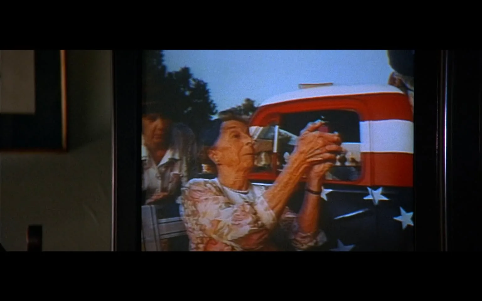The image depicts a scene from a television screen, showing two women in a somewhat rural or outdoor setting. The foreground features an elderly woman with short, dark hair, wearing a floral-patterned blouse. She appears to be holding something in her hands, possibly a small object or device, and is looking intently at it. Her expression is focused, and she has a slight smile. 

In the background, another woman is partially visible, wearing a light-colored shirt. She seems to be observing the elderly woman, but her facial expression is not clearly discernible due to the angle and focus of the shot. 

The setting includes a vintage pickup truck, which is prominently displayed on the right side of the image. The truck has a red cab and a bed decorated with a design featuring white stripes and blue stars, resembling the American flag. The background is somewhat blurred but indicates greenery, suggesting an outdoor environment, possibly a park or a rural area.

The image is framed by a dark border, likely the edges of the television screen, which adds a layer of depth to the scene. The overall lighting appears warm, suggesting it might be late afternoon or early evening. The colors are somewhat muted, typical of older film or television footage. 

No readable text, logos, or trademarks are visible in the image. The focus remains on the two women and the truck, with no additional identifiable brands or products present.