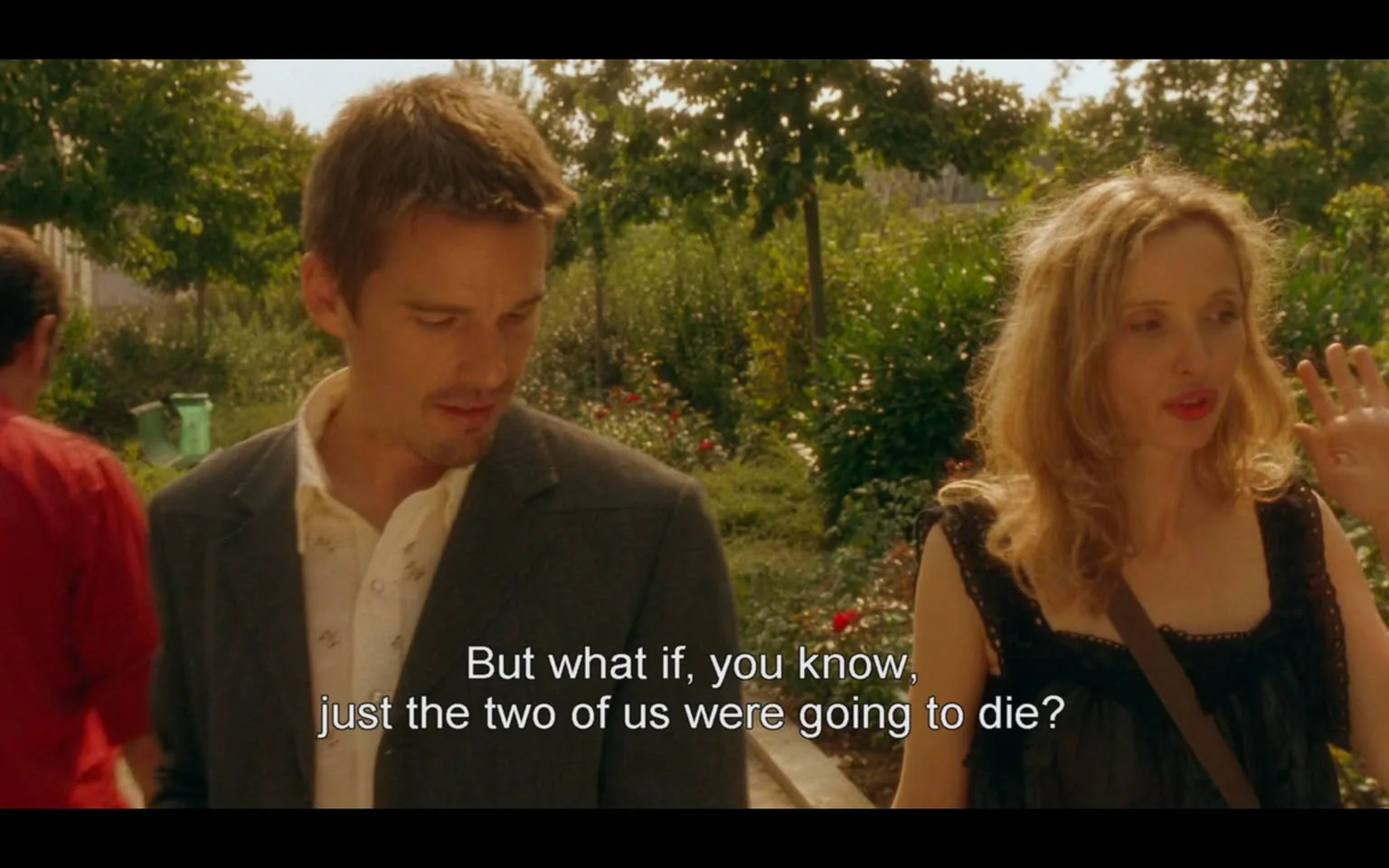 The image depicts a scene from a film featuring two characters engaged in conversation. 

**Text:** The visible text at the bottom of the image reads: "But what if, you know, just the two of us were going to die?" This text appears to be a subtitle.

**Characters:** 
1. **Male Character:** 
   - He has short, slightly tousled light brown hair and a well-groomed appearance.
   - He is wearing a dark gray suit jacket over a white shirt with a subtle pattern, which is unbuttoned at the collar.
   - His expression appears thoughtful or concerned as he looks down, not directly at the camera.

2. **Female Character:** 
   - She has long, wavy blonde hair and is wearing a black sleeveless dress with a delicate lace trim.
   - Her makeup includes bright red lipstick, and she has a neutral expression while gesturing with her right hand, possibly emphasizing a point in their conversation.
   - She appears to be looking at the male character, engaged in the discussion.

**Background:** 
- The setting is an outdoor area, likely a garden or park, with lush greenery and flowering plants visible in the background. 
- There are trees and shrubs surrounding the characters, creating a serene and natural environment.
- In the background, a person in a red shirt is partially visible, walking away from the camera.

**Lighting:** 
- The scene is brightly lit, suggesting it is daytime, with sunlight filtering through the trees, casting a warm glow on the characters.

Overall, the image captures a moment of dialogue between the two characters in a picturesque outdoor setting, with a focus on their expressions and interaction.