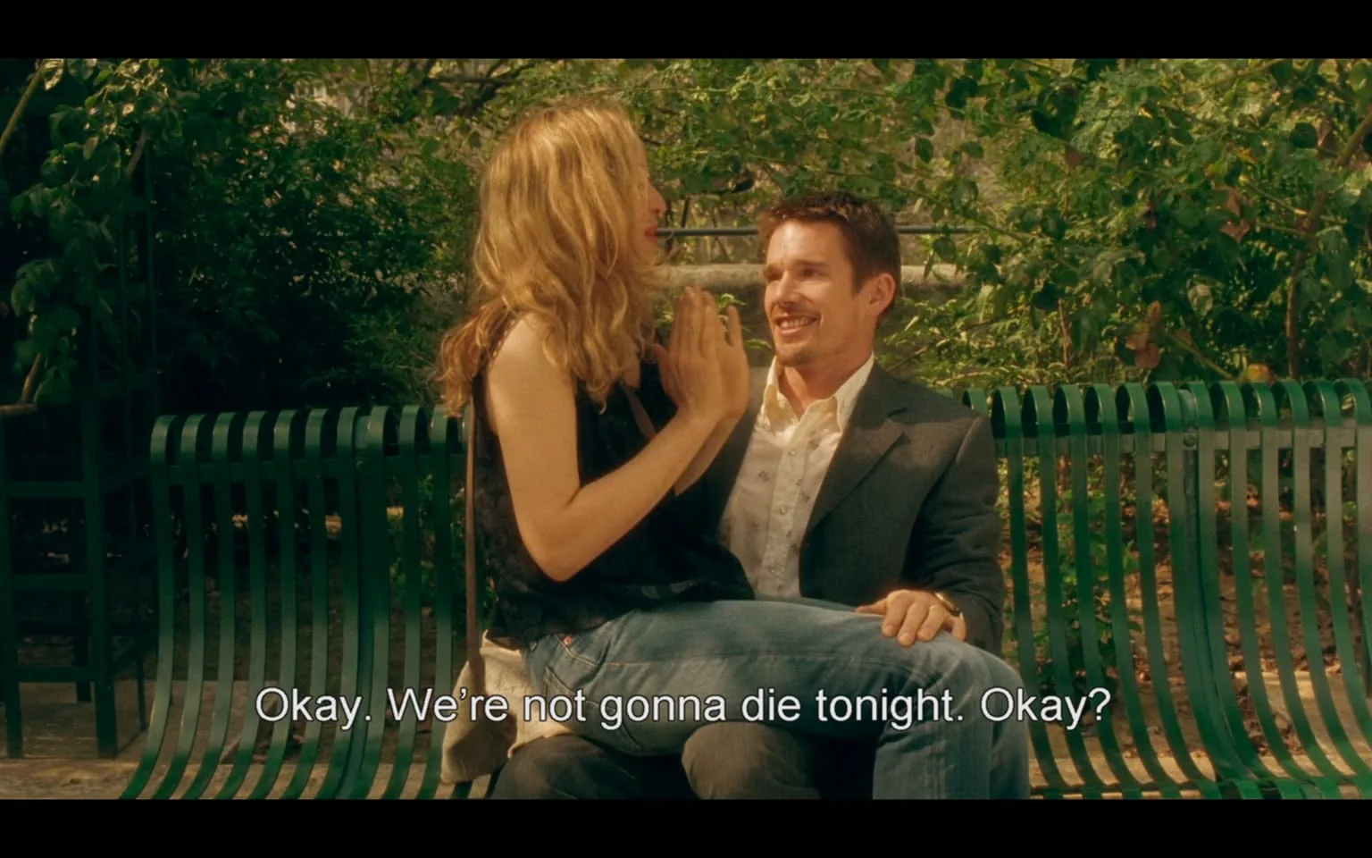 The image depicts two individuals seated on a green park bench surrounded by lush greenery. The woman on the left has long, wavy blonde hair and is wearing a black sleeveless top. She appears animated, with her hands raised in a gesture that suggests she is expressing something important. The man on the right has short, dark hair and is dressed in a light-colored shirt with a blazer. He is smiling and looking at the woman, suggesting engagement in their conversation.

The bench has a distinctive design, featuring horizontal slats painted in green, and is situated in a park-like setting with trees and foliage in the background. The sunlight filters through the leaves, creating a warm atmosphere. The text at the bottom of the image reads: "Okay. We're not gonna die tonight. Okay?" This suggests a moment of reassurance or a light-hearted exchange between the two individuals.

Overall, the scene conveys a casual and intimate moment between the two people, set in a natural environment that enhances the warmth of their interaction.