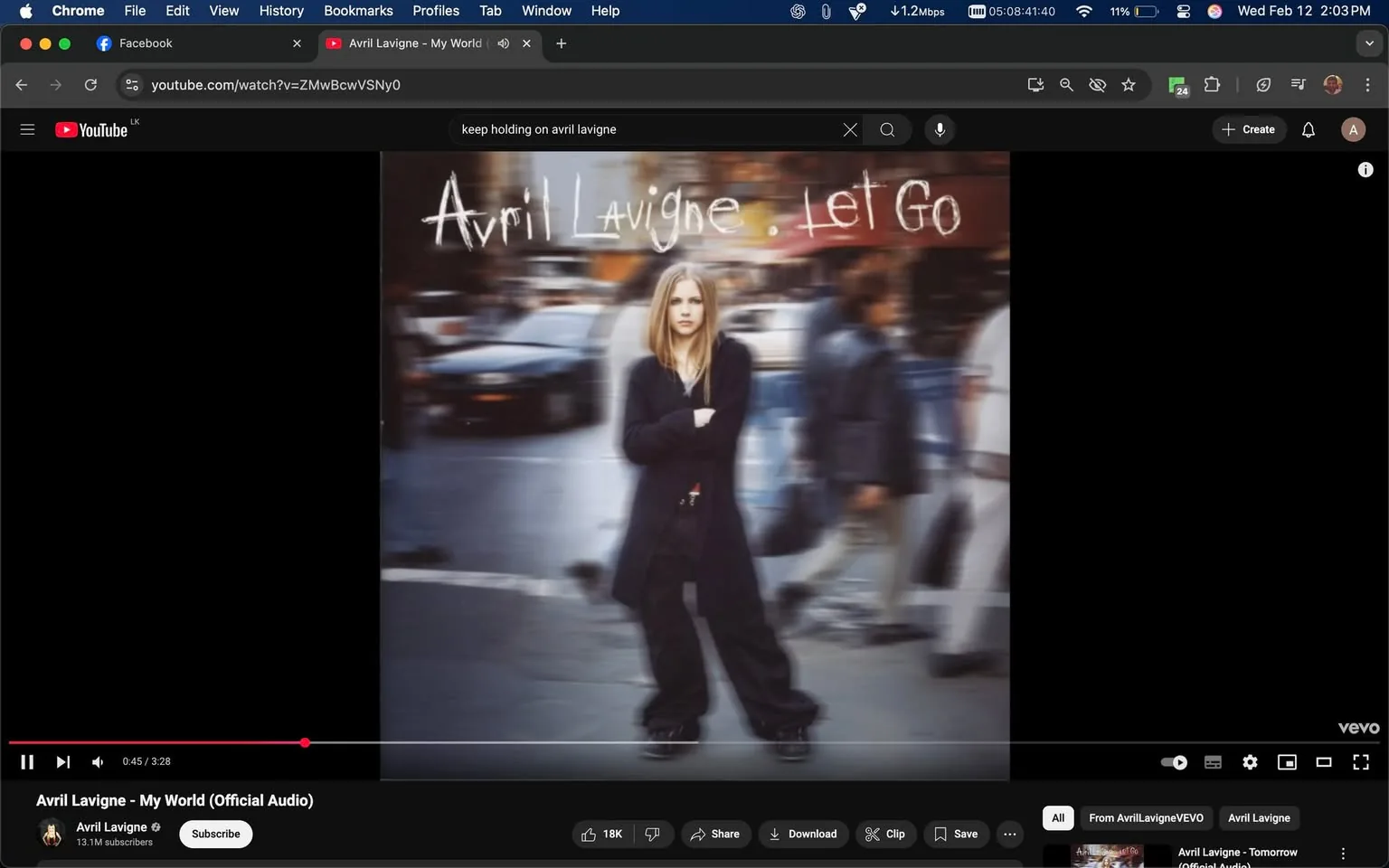 The image features an album cover for "Let Go" by Avril Lavigne. The background is a blurred urban street scene, suggesting a busy city environment with cars and pedestrians. The focus of the image is a young woman standing in the foreground, who appears to be Avril Lavigne. She has long, straight blonde hair and is wearing a long black coat that reaches her knees. Underneath the coat, she has a black top and baggy black pants, which are slightly oversized. Her footwear consists of black shoes. 

The woman is standing with her arms crossed, facing the camera, and has a neutral expression. The background shows indistinct figures of people walking, indicating movement and activity in the setting. The text "Avril Lavigne - Let Go" is prominently displayed at the top of the image in a handwritten style, with "Avril Lavigne" in a larger font size. 

The overall color palette is muted, with a focus on darker tones, which contrasts with the lighter color of the woman's hair. The image conveys a sense of urban life, with the blurred background enhancing the focus on the subject in the foreground. There are no visible logos, trademarks, or additional text apart from the album title and artist name. The image appears to be a promotional or cover art piece for the album.