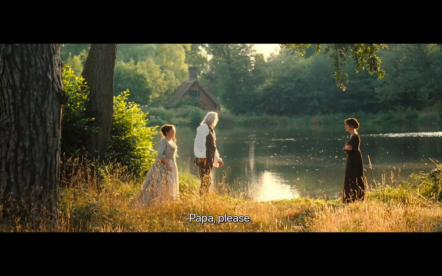 The image depicts a serene outdoor scene near a body of water, likely a pond or lake, surrounded by lush greenery. The setting is bathed in warm, golden light, suggesting either early morning or late afternoon. 

In the foreground, there are three figures:

1. **Left Figure**: A woman dressed in a light-colored, flowing gown with a floral pattern. She has dark hair styled in an updo and appears to be looking towards the other two figures. Her expression is not clearly visible due to the angle.

2. **Middle Figure**: A man with long, gray hair, wearing a white shirt and dark trousers. He stands with his back to the viewer, facing the water. He holds a small object, possibly a basket or a cloth, in his right hand. His posture suggests he is engaged in conversation with the woman beside him.

3. **Right Figure**: Another woman, dressed in a dark, fitted dress that reaches her ankles. She stands with her arms crossed, facing the water. Her hair is styled in loose waves, and she appears contemplative, looking away from the other two figures.

The background features a rustic wooden house partially obscured by trees and shrubs, indicating a rural setting. The water reflects the surrounding greenery, and there are small ripples visible on the surface. The area is overgrown with tall grass and wildflowers, contributing to the natural ambiance.

No readable text, logos, or brands are present in the image. The overall composition conveys a peaceful, pastoral scene without any identifiable commercial elements.