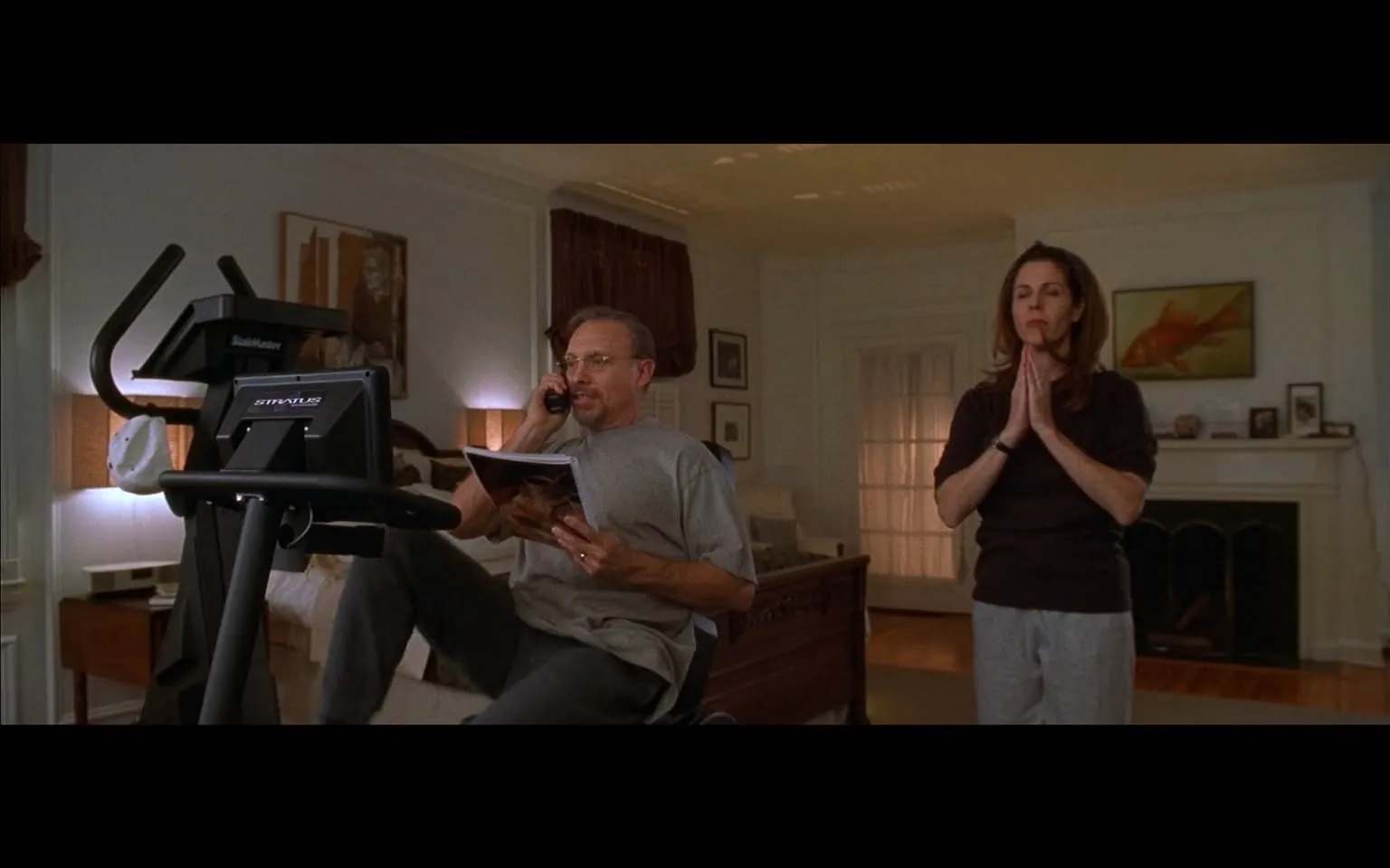 The image depicts a domestic setting with two individuals engaged in different activities. 

In the foreground, a man is seated on an exercise bike, appearing to be in the middle of a phone conversation. He has short, light brown hair and wears glasses. His attire consists of a gray short-sleeved t-shirt and dark pants. He holds a magazine in his left hand while using his right hand to hold the phone to his ear. The exercise bike is a model labeled "StairMaster," which is positioned in front of a wall adorned with framed pictures, including one that appears to depict a city skyline.

In the background, a woman stands with her hands pressed together in a prayer-like gesture. She has long, dark hair and wears a dark sweater and light gray sweatpants. Her expression is serious, and she appears to be focused on the man. The setting includes a well-furnished living room with a wooden bed frame visible to the left and a fireplace with a decorative mantle in the background. The walls are painted a light color, and there are curtains drawn at the windows, allowing some light to filter in.

The overall atmosphere of the room is calm and domestic, with warm lighting coming from lamps placed around the space. The decor includes various framed artworks and a large painting of a fish hanging above the fireplace. The floor is wooden, and the room appears tidy and organized.