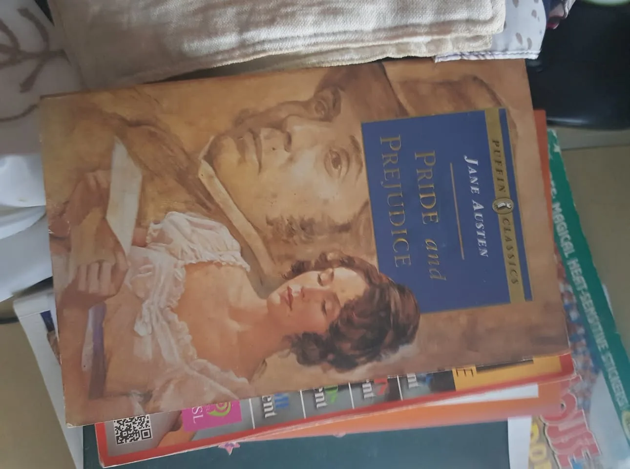 The image features a book titled "Pride and Prejudice" by Jane Austen, prominently displayed on top of a stack of other items. The cover art depicts a woman in period attire, seemingly lost in thought, with a faint image of a man in the background. The book is published under the Puffin Classics series, indicating its status as a classic literary work. Surrounding the book are various magazines and other reading materials, suggesting a casual and cozy reading environment. The setting appears to be a home or personal space, with soft lighting and a hint of fabric in the background, adding to the intimate atmosphere. There are no visible people in the image, and the content is entirely focused on the book and its surroundings, making it suitable for all audiences. The overall composition conveys a sense of literary appreciation and a love for reading.