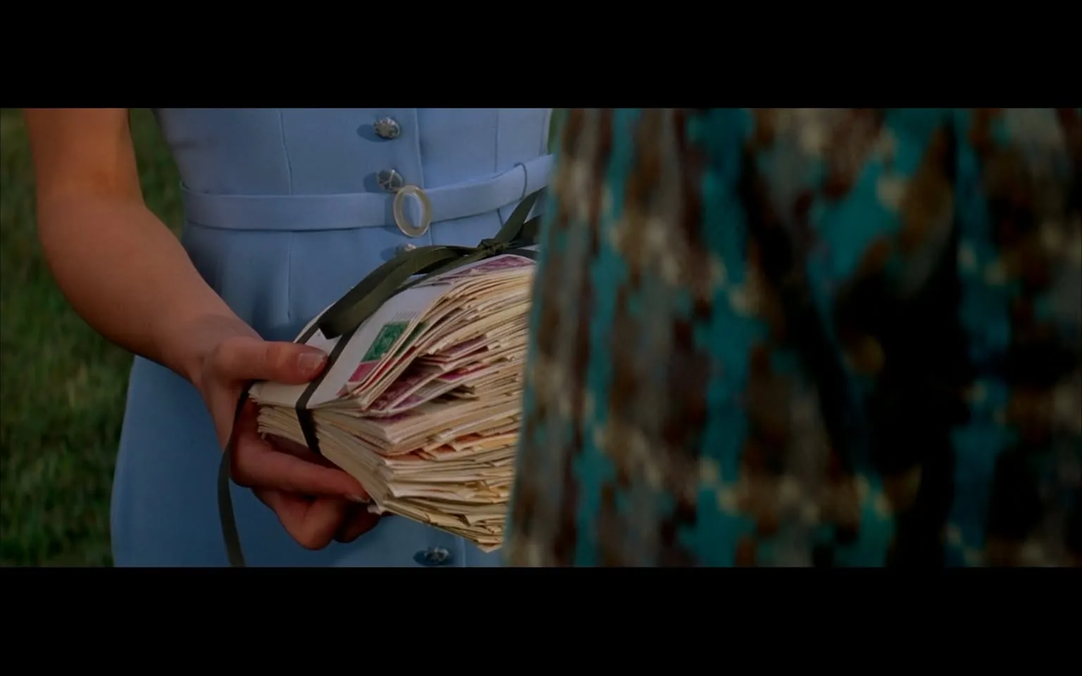 The image depicts a close-up scene featuring two individuals, with a focus on one person's hands holding a large bundle of letters. 

**Observable Details:**

1. **Letters:** The bundle consists of numerous envelopes, some of which are partially visible. The envelopes appear to be various colors, with at least one envelope showing a green stamp. The letters are tied together with a dark green ribbon.

2. **Clothing:**
   - The person holding the letters is wearing a light blue dress. The dress has a fitted bodice with a round neckline and short sleeves. There are several buttons visible down the front of the dress, and a belt cinches the waist, featuring a circular buckle.
   - The second individual, who is partially visible in the foreground, is wearing a patterned garment that appears to be a blouse or dress. The fabric features a mix of colors, including teal, brown, and cream, with a textured pattern that is not clearly defined.

3. **Hands and Appearance:** 
   - The hands of the person holding the letters are delicate, with fingers gently gripping the bundle. The nails are not prominently visible.
   - The second person's hands are also visible, suggesting a relaxed grip or gesture, but specific details about their hands are not discernible.

4. **Setting:** 
   - The background is blurred, but it suggests an outdoor setting, likely a grassy area, as indicated by the green color visible behind the individuals. The lighting appears natural, possibly indicating daytime.

5. **Facial Expressions:** 
   - The facial expressions of both individuals are not visible in this image, as the focus is primarily on the hands and the letters.

Overall, the image captures a moment of interaction involving a significant number of letters being presented, with a clear emphasis on the clothing and the bundle of correspondence. The setting appears to be outdoors, contributing to the overall context of the scene.