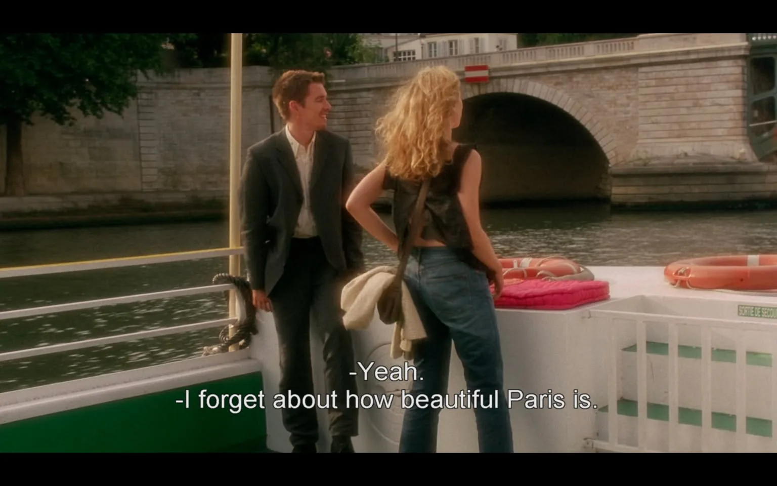 The image depicts a scene on a boat, likely on a river in Paris, given the context of the dialogue. 

**Setting and Background:**
- The background features a stone bridge with an arch, typical of Parisian architecture. The bridge has a light-colored stone facade and is adorned with a red and white sign, possibly indicating a no-entry or warning sign.
- The water is calm, reflecting the surrounding greenery and the bridge. There are trees lining the riverbank, adding to the scenic view.

**Visible Objects:**
- On the boat, there are two life rings, one orange and white, placed on the side of the boat. There is also a pink cushion or mat on the boat's deck.
- The boat itself has a green and white color scheme, with a railing visible in the foreground.

**People:**
- There are two individuals in the foreground:
  1. **Person on the Left:**
     - Male, wearing a dark suit with a white shirt. He has short, neatly styled hair and is smiling, appearing engaged in conversation.
     - His posture is relaxed, standing with his hands in his pockets.
  2. **Person on the Right:**
     - Female, wearing a sleeveless black top and blue jeans. Her hair is long and wavy, cascading down her shoulders.
     - She is facing the male, with a slight smile, suggesting a friendly interaction. She has a bag slung over her shoulder, which appears to be light-colored.

**Text:**
- The dialogue displayed on the screen reads:
  - "-Yeah."
  - "-I forget about how beautiful Paris is."
  
This text indicates a conversation taking place between the two individuals, reflecting on the beauty of Paris.

Overall, the image captures a moment of casual conversation between two people on a boat, set against a picturesque Parisian backdrop.