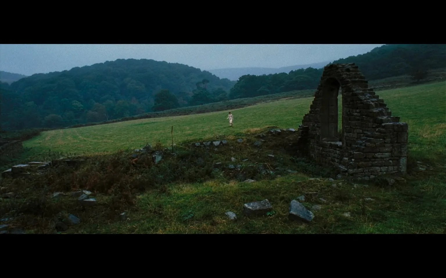 The image depicts a serene landscape characterized by a grassy field and a partially ruined stone structure on the right side. The structure appears to be an old stone wall with an arched opening, suggesting it may have once been part of a building, possibly a chapel or barn. The stones are irregularly shaped and weathered, indicating age and exposure to the elements. 

In the foreground, there are scattered stones and patches of grass, with some areas showing signs of overgrowth, including brownish and green vegetation. The ground is uneven, with tufts of grass and small rocks visible.

In the background, rolling hills are visible, covered with dense trees that display various shades of green, indicating a lush environment. The sky is overcast, with a grayish hue, suggesting a cloudy or possibly rainy day. The lighting is soft, contributing to a tranquil atmosphere.

A figure is present in the field, located towards the center-left of the image. The person is wearing a light-colored, flowing garment, which appears to be a dress or long shirt. The figure is walking away from the camera, with their back turned, and their hair is not clearly visible. The posture does not convey any specific emotion or action beyond walking.

Overall, the scene is peaceful and natural, with no visible text, signs, or identifiable brands. The focus is on the landscape and the solitary figure within it.