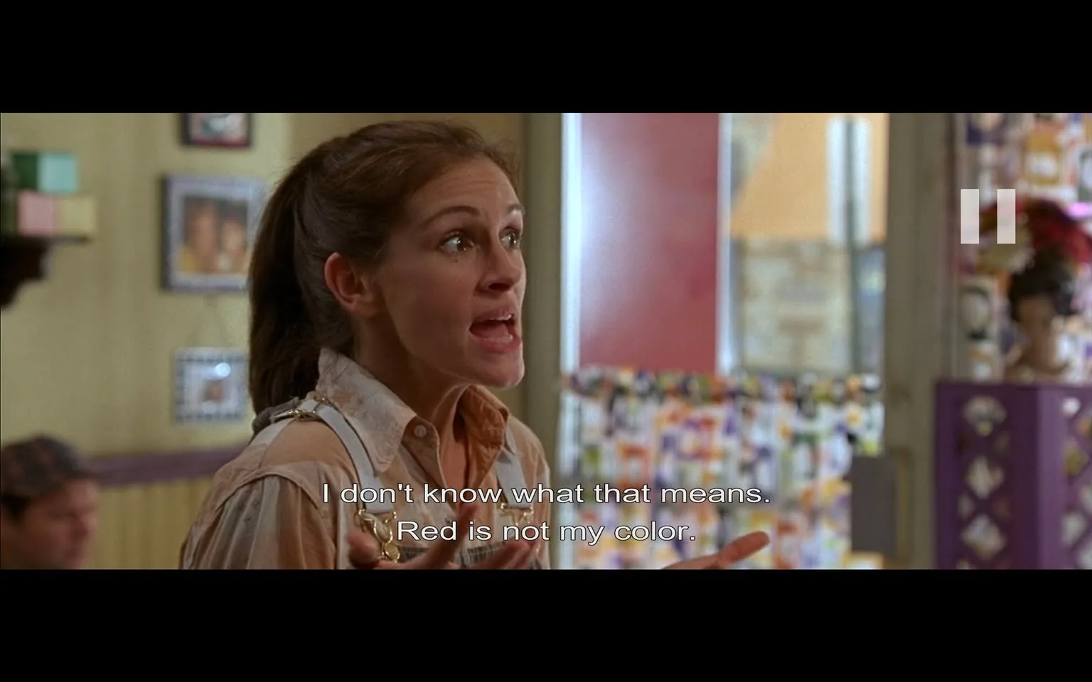 The image depicts a scene from a film featuring a woman who appears to be in a state of agitation or frustration. She has long, brown hair pulled back and is wearing a light-colored, button-up shirt with a collar. The shirt has a slightly wrinkled appearance, and she is gesturing with her hands as she speaks, indicating a strong emotional response. Her facial expression shows intensity, with wide eyes and slightly parted lips, suggesting she is in the midst of a passionate dialogue.

In the background, there are various decorative elements typical of a casual dining or retail environment. The walls are adorned with framed photographs and colorful decorations, creating a lively atmosphere. A counter or display area is visible, with a blurred figure in a cap seated in the background, possibly indicating a customer or employee. The setting appears to be a small, informal establishment, likely a café or diner, given the casual decor and the presence of food-related items.

The text displayed on the screen reads: "I don't know what that means. Red is not my color." This suggests that the woman is responding to a comment or suggestion regarding color, possibly related to clothing or style. The overall composition captures a moment of dialogue that conveys a sense of urgency or disagreement.

No identifiable brands, logos, or trademarks are visible in the image. The focus remains on the woman and her expression, with the background serving to enhance the context of the scene.