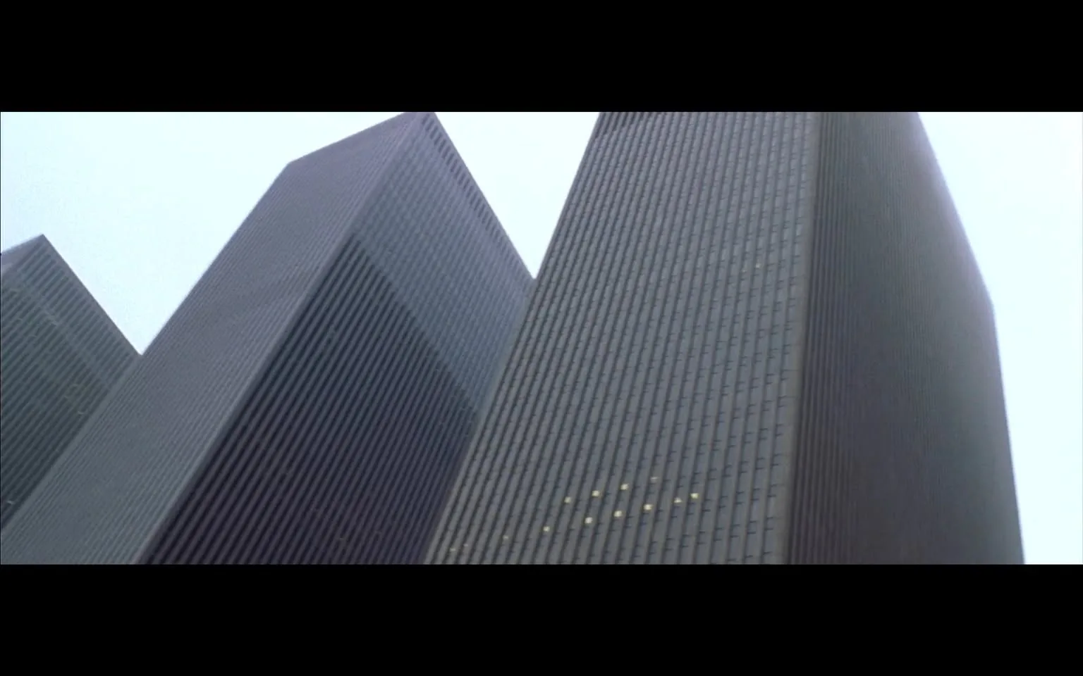 The image depicts two tall, modern skyscrapers, characterized by their sleek, glass and steel facades. The buildings are primarily gray and feature vertical lines created by the arrangement of their windows, which are arranged in a grid pattern. The left building has a slightly angled top, while the right building has a more traditional rectangular shape, both contributing to a contemporary architectural style.

The sky above is overcast, suggesting a cloudy day, which adds a muted tone to the overall scene. The buildings appear to be part of an urban environment, likely in a city center, given their height and design. There are no visible signs, logos, or text on the buildings themselves, and the image does not include any people or vehicles, focusing solely on the architectural elements.

The perspective of the photograph is from below, looking up at the buildings, which emphasizes their height and grandeur. The lower part of the image is darker, possibly due to shadows cast by the buildings, while the upper part is lighter, where the sky is visible. Overall, the image conveys a sense of modernity and urban life, with a focus on the architectural features of the skyscrapers.
