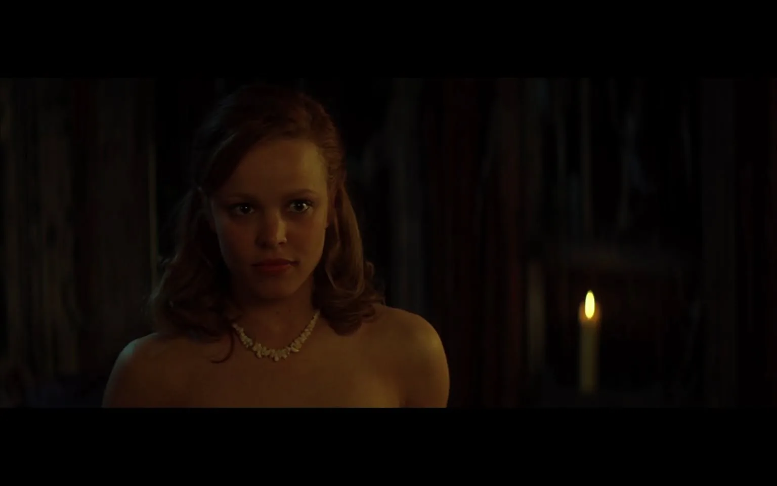 The image features a young woman standing in a dimly lit environment. She has long, wavy hair that appears to be a light brown or auburn color. Her hair is styled with soft curls, framing her face. The woman is wearing a strapless dress, which is not clearly visible in detail due to the lighting, but it appears to be a light color, possibly beige or pale pink. Around her neck, she is adorned with a delicate necklace that consists of small, round beads, adding a touch of elegance to her appearance.

Her facial expression is serious and focused, with her lips slightly parted. She has a fair complexion, and her makeup is subtle, enhancing her features without being overly dramatic. The background is dark, with indistinct shapes and shadows, suggesting a setting that is intimate or possibly mysterious. There is a single candle in the background, emitting a soft, warm glow, which highlights her face and creates a contrast with the surrounding darkness.

No readable text, logos, or brands are present in the image. The overall atmosphere of the scene is one of tension or anticipation, but specific emotions or relationships cannot be inferred from the visual details alone. The setting does not provide clear indicators of a specific event or location, but the ambiance suggests a private or personal moment.