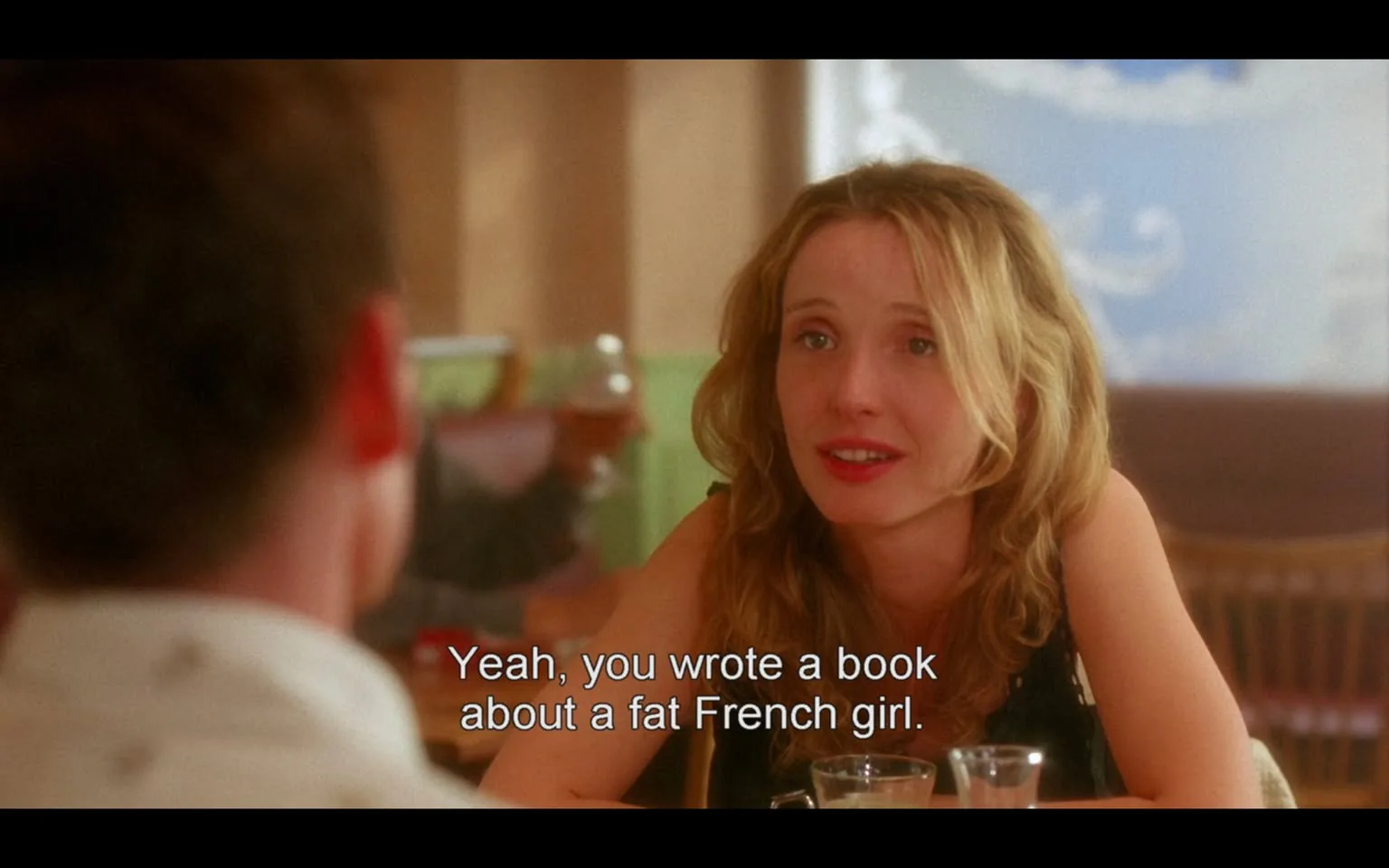 The image depicts a scene from a film, featuring a woman engaged in conversation with a man, who is partially out of frame. 

**Visible Text:**
- The subtitle reads: "Yeah, you wrote a book about a fat French girl."

**People:**
- The woman has long, wavy blonde hair and is wearing a black sleeveless top. She has a light complexion and appears to be smiling or engaged in a friendly conversation. Her facial expression suggests interest or amusement.
- The man, whose face is not fully visible, is dressed in a light-colored shirt. His head is turned slightly away from the camera, making it difficult to discern his expression.

**Setting:**
- The background features a restaurant or café setting, with soft lighting and a warm color palette. There are wooden chairs and tables visible, indicating a casual dining environment.
- In the background, there is a green wall with some decorative elements, possibly paintings or murals, that add to the ambiance of the place. A glass of beverage is visible on the table in front of the woman, along with another glass, suggesting they are in the midst of a meal or drinks.

**Overall Context:**
- The scene captures a moment of dialogue between the two characters, likely during a casual meeting or date. The atmosphere appears relaxed and informal, typical of a café setting. 

This description focuses on observable details without inferring emotions or relationships.