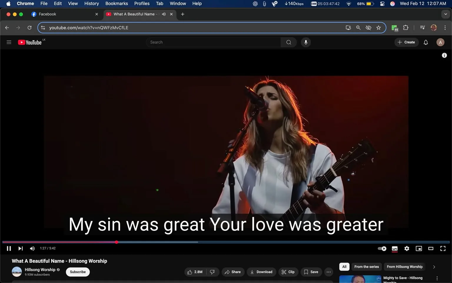 The image shows a scene from a YouTube video titled "What A Beautiful Name - Hillsong Worship." The video features a female singer performing on stage. She has long, wavy hair that is partially illuminated by stage lights, giving it a warm glow. The singer is wearing a loose-fitting white top and is holding an acoustic guitar. She appears to be singing into a microphone, with her eyes closed, suggesting a moment of emotional engagement with the performance.

The background is dark, with a gradient of red and orange lighting that creates a dramatic atmosphere. The lighting highlights the singer's face and hair, while the rest of the stage remains dimly lit. Below the singer, there are subtitles displaying the lyrics: "My sin was great Your love was greater," which are clearly visible in white text against a dark background.

In the top portion of the image, the YouTube interface is visible, including the video title, channel name "Hillsong Worship," and the number of subscribers (9.93M). There are also playback controls at the bottom, showing the current time of the video (1:27) and the total duration (5:42). The video has a view count of 2.8 million, indicating its popularity.

The overall setting appears to be a live worship performance, likely in a church or concert venue, characterized by the intimate and reverent atmosphere typical of such events. The focus is on the singer and her performance, with no other performers or audience members visible in the frame.