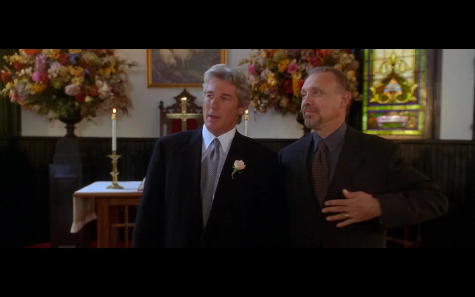 The image depicts two men standing inside a church setting. 

**Setting and Background:**
- The interior of the church features a dark wooden paneling, with a red carpet on the floor.
- In the background, there is a large floral arrangement consisting of various flowers in shades of pink, yellow, and white, placed in a tall vase.
- To the left, there is a lit candle on a golden candlestick, and behind it, a wooden altar with a red cloth draped over it.
- A stained glass window is visible on the right side of the image, featuring intricate designs and colors, primarily in green and blue hues.
- There is a wooden cross on the altar, indicating the religious context of the setting.

**People:**
1. **First Man:**
   - He has light gray hair and is wearing a black suit with a white dress shirt underneath.
   - A light pink rose is pinned to his lapel.
   - His facial expression appears neutral, and he is looking slightly to his left.
   - He has a slight smile and is gesturing with his hands, possibly speaking or reacting to something.

2. **Second Man:**
   - He has a darker complexion and is dressed in a dark suit with a light blue dress shirt and a patterned tie.
   - His hair is short and neatly styled, and he has a well-groomed beard.
   - He is looking at the first man, with a slight smile on his face, and his right hand is raised, possibly gesturing or emphasizing a point.

**Clothing Details:**
- Both men are dressed formally, indicating that this is likely a special occasion, such as a wedding or a religious ceremony.
- The first man’s suit is classic and tailored, while the second man’s suit appears slightly more modern with a patterned tie.

Overall, the image captures a moment in a formal setting, likely during a significant event, with two men engaged in conversation or interaction.