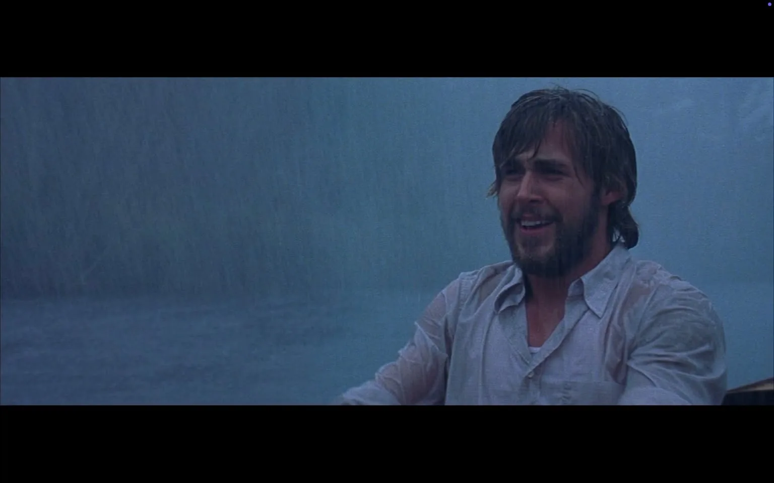 The image depicts a man in a rain-soaked environment, suggesting a stormy or heavy rain setting. He has a medium build and appears to be in a boat or on a body of water. His hair is medium-length, slightly disheveled, and appears wet, indicating exposure to rain. He has a beard and mustache, both of which are also wet. 

The man is wearing a light-colored, button-up shirt that is soaked through, clinging to his body. The shirt has a collar and appears to be unbuttoned at the top. His facial expression shows a wide smile, and he seems to be laughing or expressing joy, despite the adverse weather conditions. 

The background is dominated by a heavy downpour, with rain visibly cascading down, creating a blurred effect in the air. The water surface appears disturbed, likely due to the rain, and the overall color palette is muted, with shades of blue and gray dominating the scene. 

There are no readable texts, logos, or identifiable brands in the image. The focus is solely on the man and the natural elements surrounding him. The setting suggests an outdoor environment, possibly a lake or river, given the presence of water and the absence of any urban structures. The overall atmosphere conveys a sense of adventure or spontaneity in the face of challenging weather.