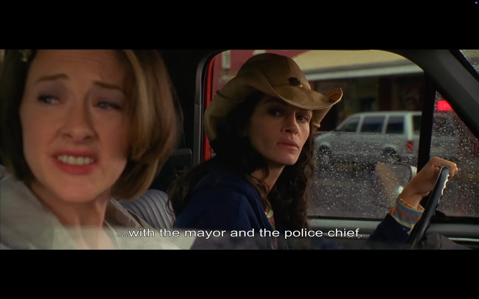 The image features two women seated inside a vehicle, likely a truck, with rain visible on the windows, suggesting an overcast or rainy day. 

**Left Woman:**
- She has medium-length, straight brown hair and is wearing a light-colored, possibly white or beige, top. 
- Her facial expression appears concerned or distressed, with her eyebrows slightly furrowed and her mouth turned down, indicating discomfort or disapproval.
- She is looking towards the right, away from the camera.

**Right Woman:**
- She has long, dark hair styled in loose waves and is wearing a dark blue sweater. 
- On her head, she sports a light-colored cowboy hat, which has a wide brim and a decorative band.
- Her expression is serious and focused, as she looks straight ahead, possibly at the road or something outside the vehicle.
- Her right hand is on the steering wheel, indicating she is driving.

**Background:**
- The interior of the vehicle is visible, with a gray seat and a red panel or dashboard.
- Outside the vehicle, blurred shapes of cars and buildings can be seen, suggesting a small-town setting with shops or businesses in the background. The rain on the window adds a layer of texture to the scene.

**Text:**
- At the bottom of the image, there is a subtitle that reads, "...with the mayor and the police chief," indicating a conversation taking place between the two women.

Overall, the image captures a moment of dialogue between the two women, set against a rainy backdrop, with a focus on their expressions and the vehicle's interior.