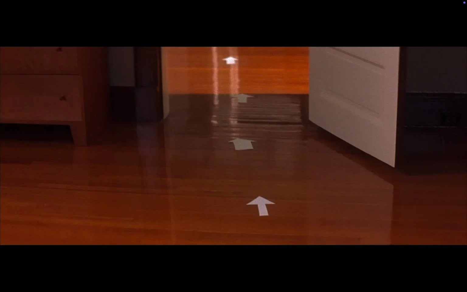 The image depicts a wooden floor with a glossy finish, reflecting light. The floorboards are arranged in a linear pattern, showcasing a rich, warm brown color. In the foreground, there is a set of white arrows made from paper or adhesive material, pointing towards an open door. The arrows are evenly spaced and lead in a straight line, indicating a direction.

To the left, there is a piece of furniture that appears to be a wooden nightstand or dresser. It has a simple design with a smooth surface and a few visible drawers. The wood grain is visible, adding texture to the piece. The edges of the furniture are rounded, and it is positioned close to the wall.

In the background, there is an open door that leads into another room. The door is white and has a simple design with no visible embellishments. The wall adjacent to the door is painted in a darker color, contrasting with the light-colored door. The floor continues into the room beyond the door, maintaining the same wooden finish.

The overall setting appears to be an indoor space, likely a home or a similar environment, characterized by its clean and organized appearance. There are no people present in the image, and no additional objects or decorations are visible in the immediate vicinity. The lighting is warm and inviting, enhancing the wooden tones of the floor and furniture.